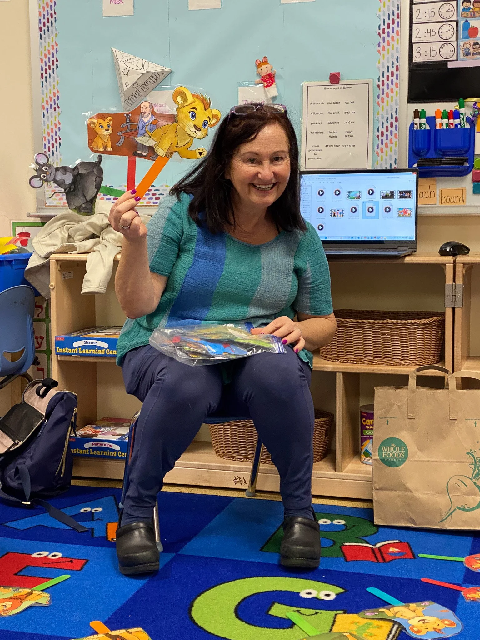 A woman sitting on a chair in a colorful classroom, holding a puppeteering stick with lion, mouse, and other animal puppets. She is smiling, and there are educational posters, a computer, and Montessori materials in the background.