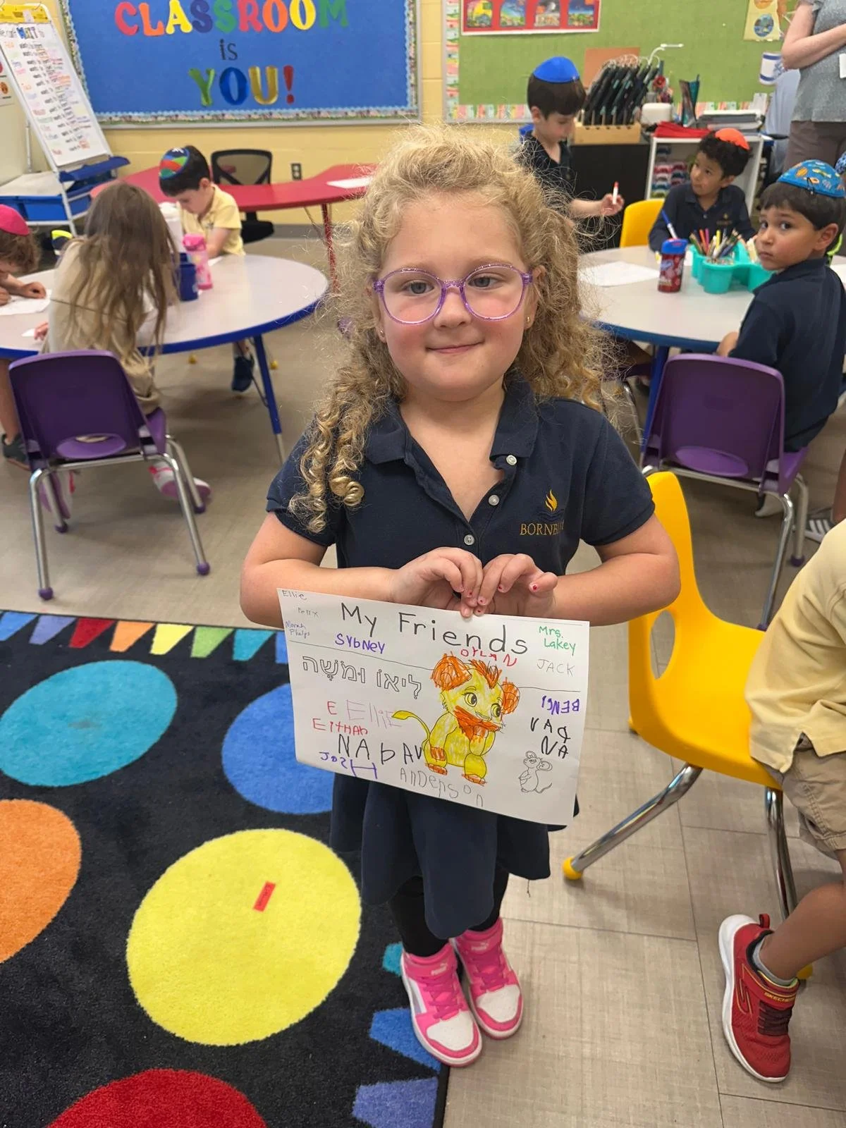 A young girl with curly blonde hair and pink glasses stands in a colorful classroom, holding a drawing with the title 'My Friends' and names of friends written around a cute lion drawing. Other children are sitting at tables in the background, some w