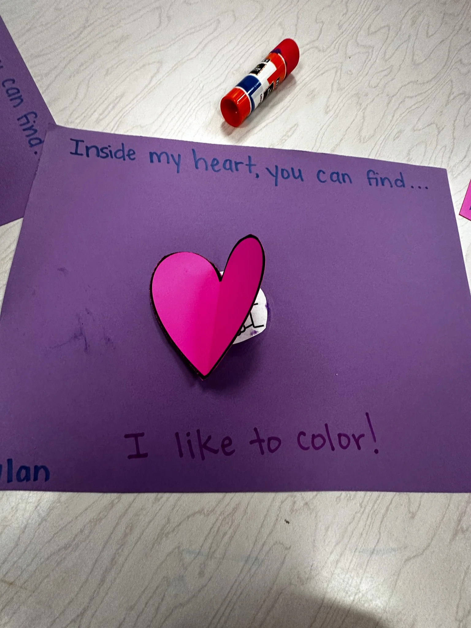 A purple sheet of paper with a cutout pink heart attached to it, containing handwriting that says, 'Inside my heart, you can find... I like to color!'. A glue stick is lying on the table above the paper.