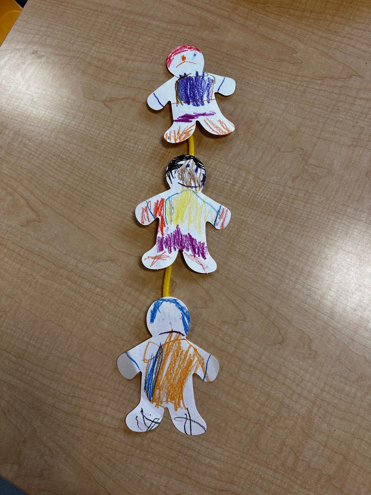Three paper cutouts of people, drawn and colored with crayons, connected by a yellow pipe cleaner on a wooden surface.