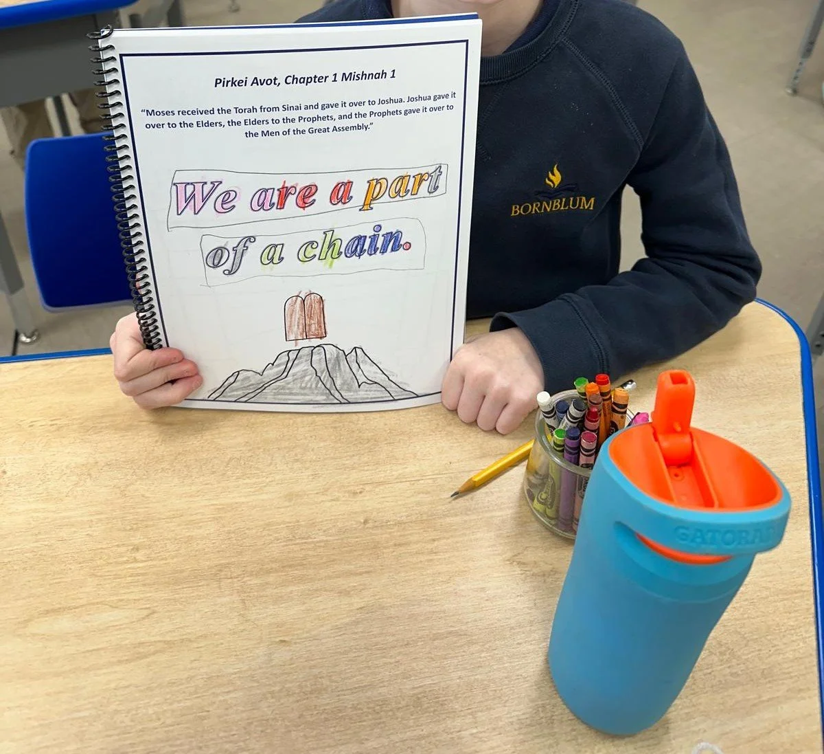 A child holding a notebook with a Bible verse and colorful handwriting, seated at a desk with a container of markers, a yellow pencil, and a blue water bottle in a classroom setting.