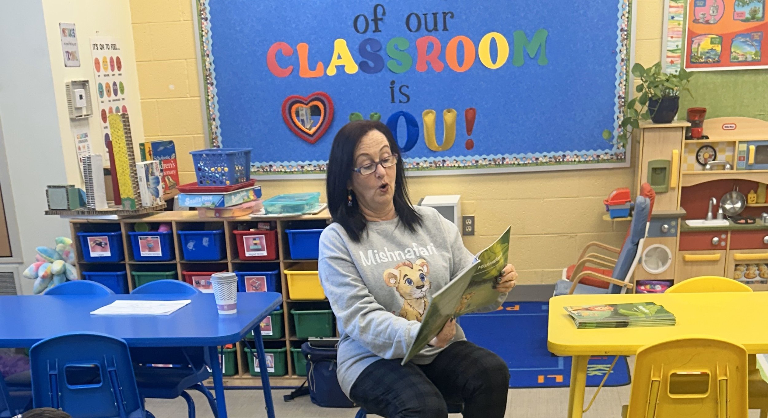 A woman reading a children's book to a classroom, with colorful tables and storage bins, a bulletin board that says "of our classroom is YOU!", and a small play kitchen area in the background.