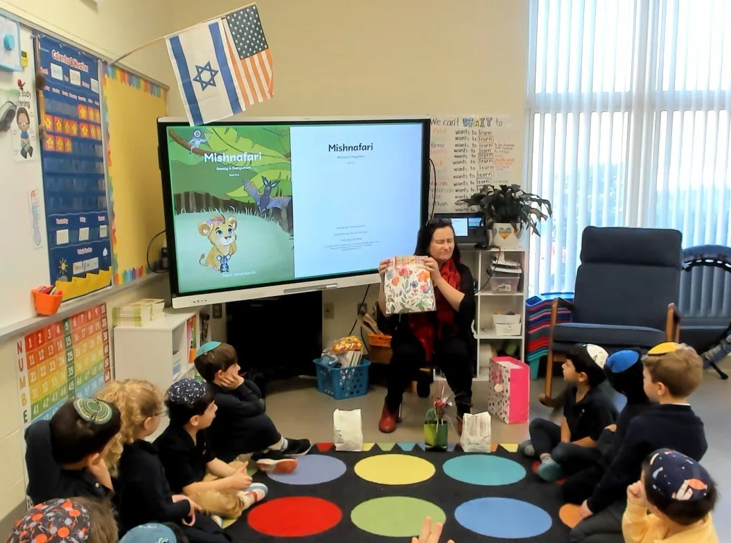 A teacher reading a story to a group of young children in a classroom. The teacher is holding a book and sitting in front of a large screen displaying a children's book titled 'Mishnafari.' The children are sitting on a colorful polka dot rug, wearin