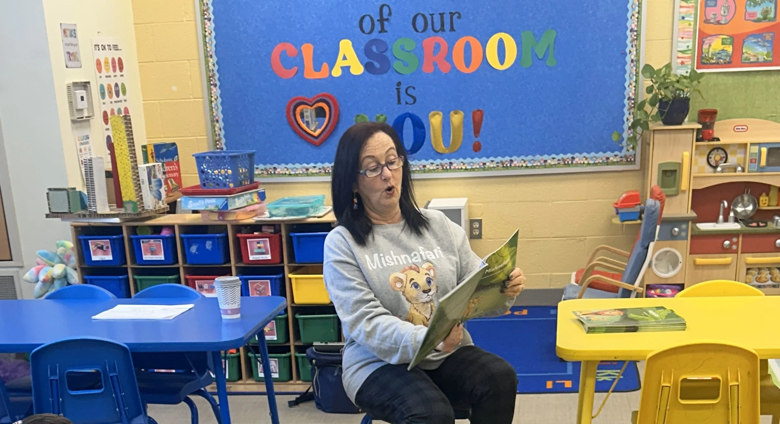 A woman sitting in a classroom reading a book, with colorful tables and cubbies behind her. The classroom has a blue bulletin board that says 'of our classroom is you!' with colorful letters and decorations.