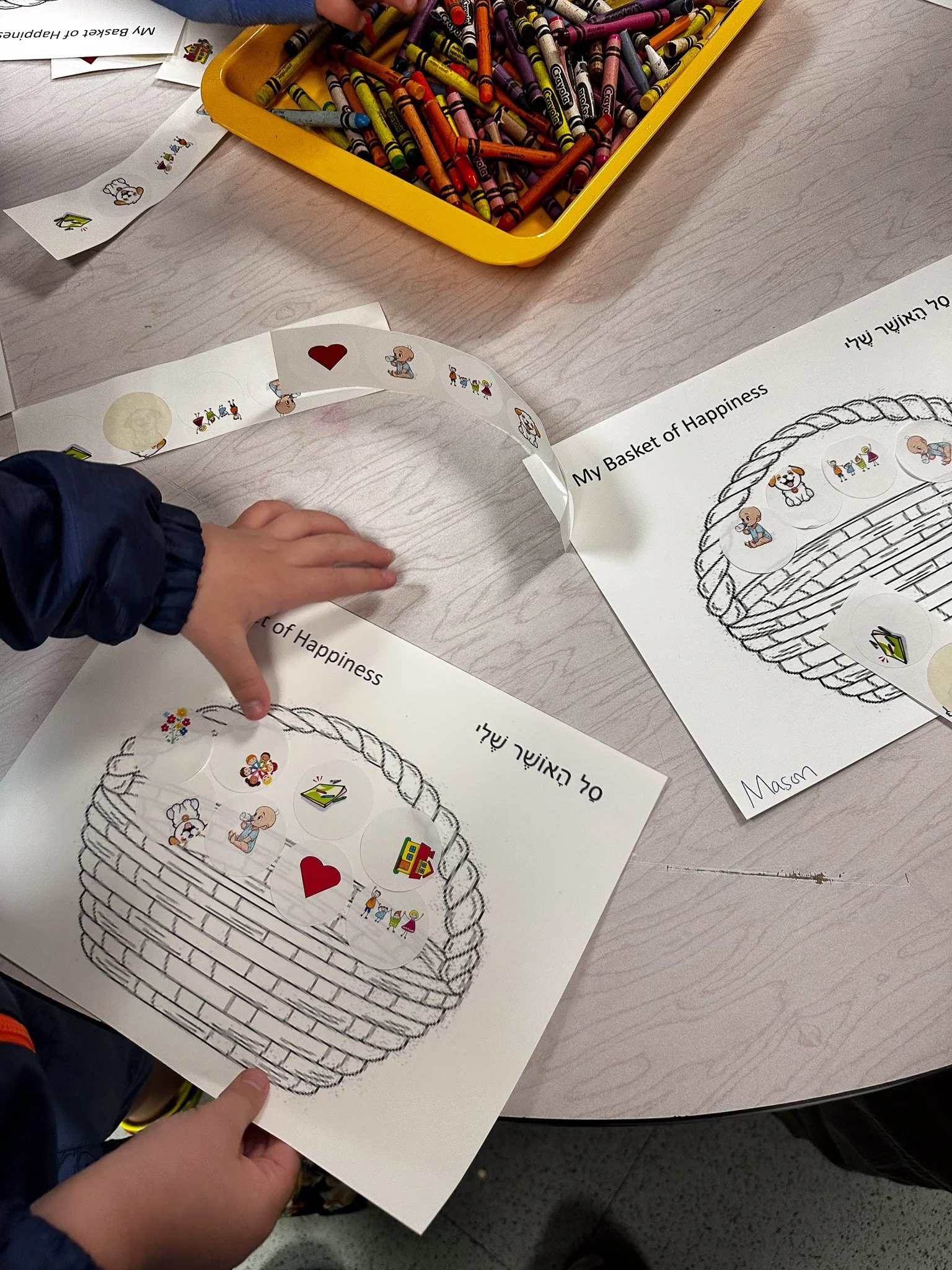 Child placing stickers on a worksheet titled 'My Basket of Happiness', which features a large basket drawing with circular stickers of various images, including a heart, a doctor, a house, a family, a cake, a puppy, and a teddy bear. There is a yello