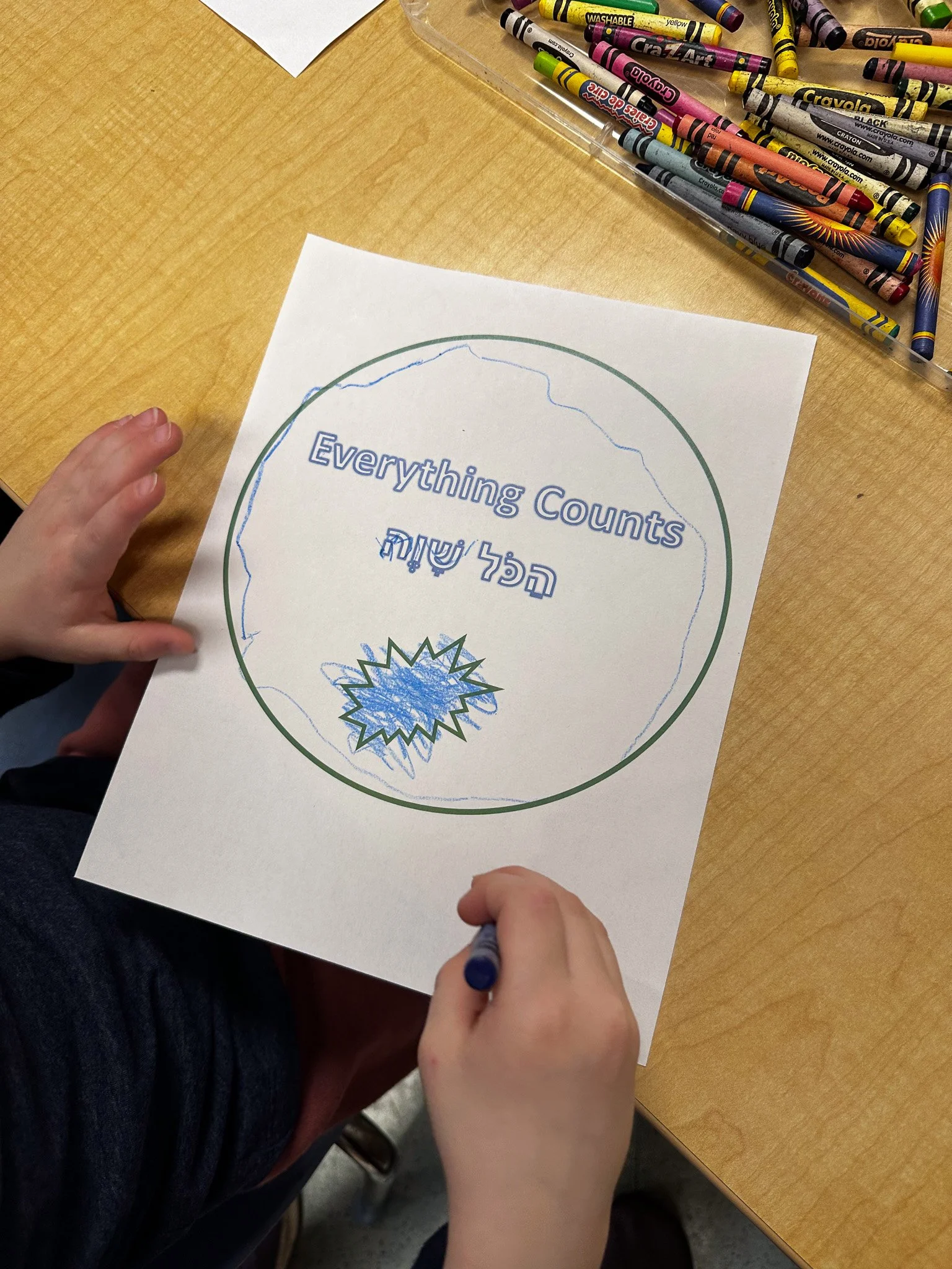 A child is writing on a sheet of paper with the phrase "Everything Counts" and Hebrew translation inside a circle, resting on a wooden table with a box of colored crayons nearby.