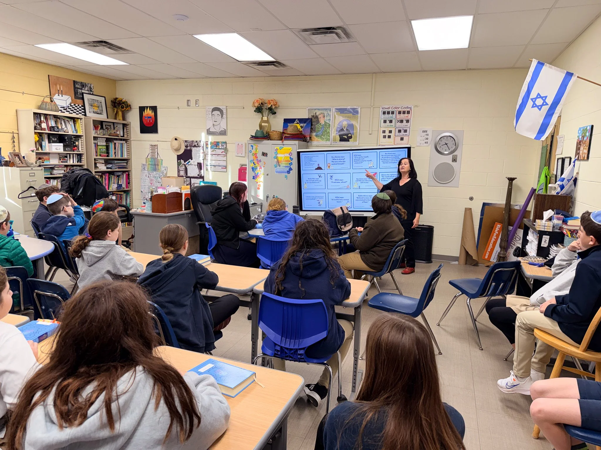A classroom with students seated at desks, listening to a female teacher standing in front of a digital display. The classroom is decorated with posters, bookshelves, and an Israeli flag hanging on the wall.