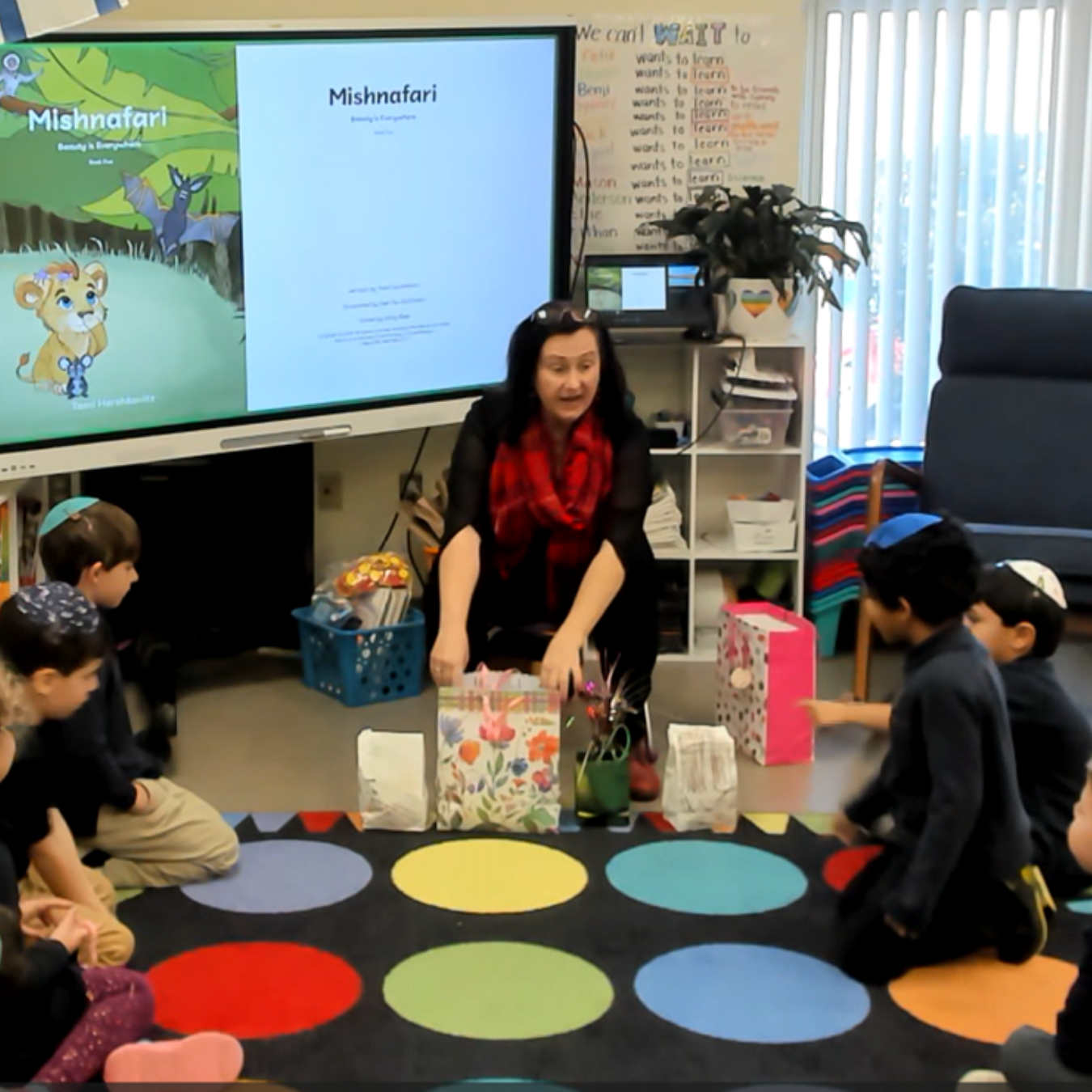 A woman leading a classroom activity with children sitting on a colorful polka dot carpet, gift bags in front of her, in a room with a large screen displaying the cover of a book titled 'Mishnafari.'
