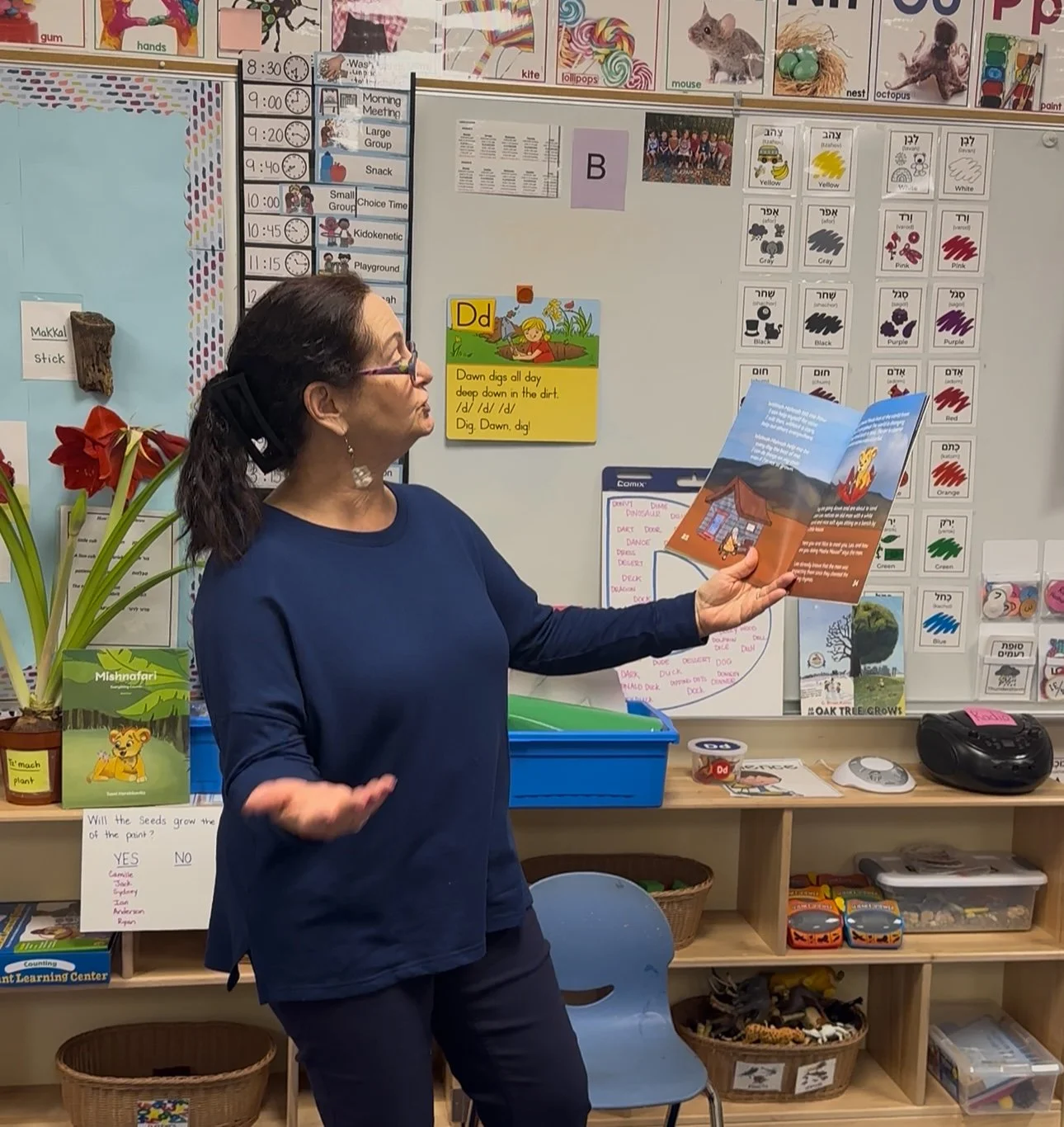 A woman with dark hair tied back, wearing glasses and a navy blue long-sleeve shirt, stands in a classroom holding an open picture book, reading aloud to children. The classroom has educational posters, a calendar, and craft supplies on shelves and a