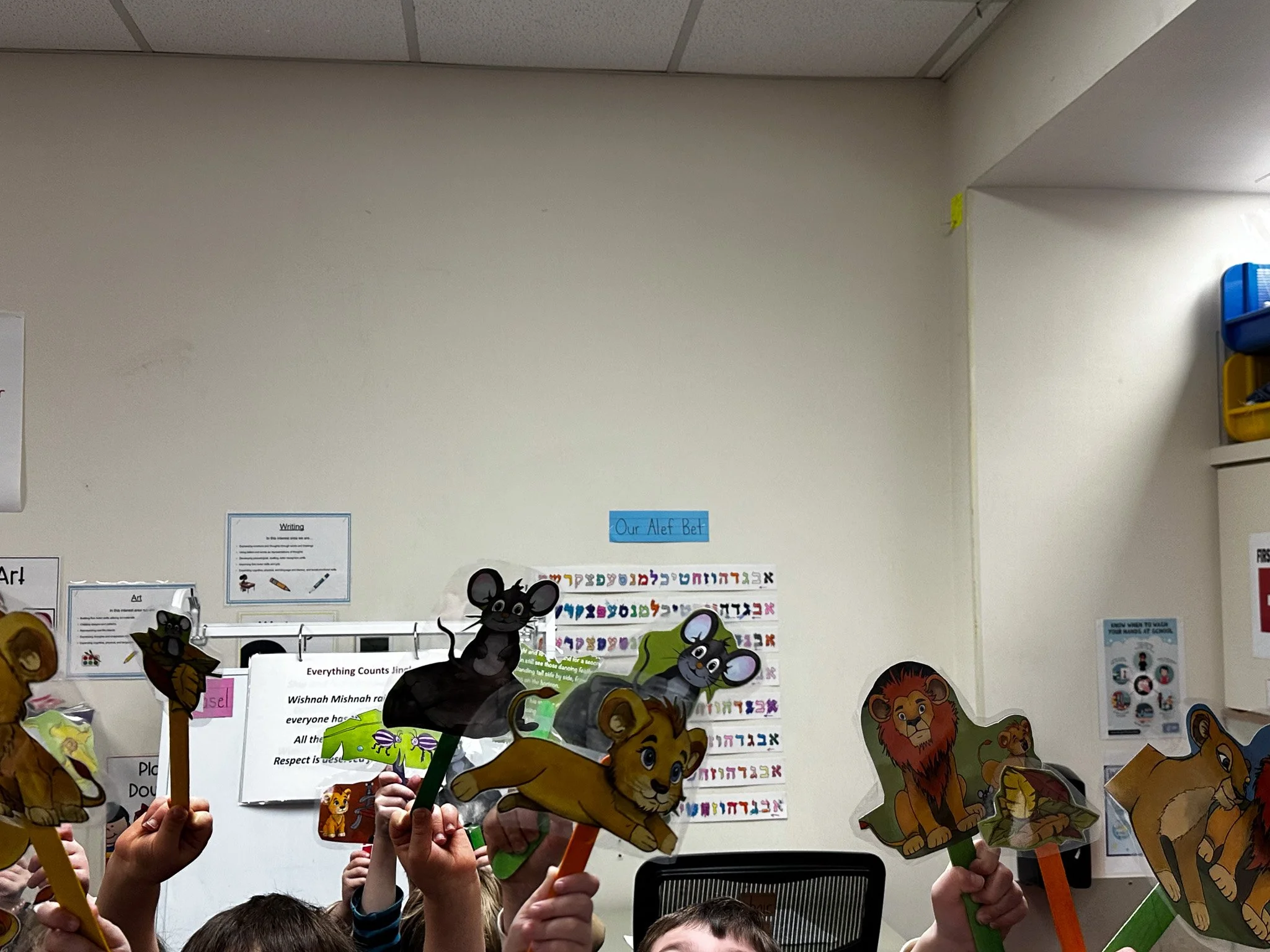 Children in a classroom holding animal puppets, including lions, mice, and a zebra, raised above their heads.