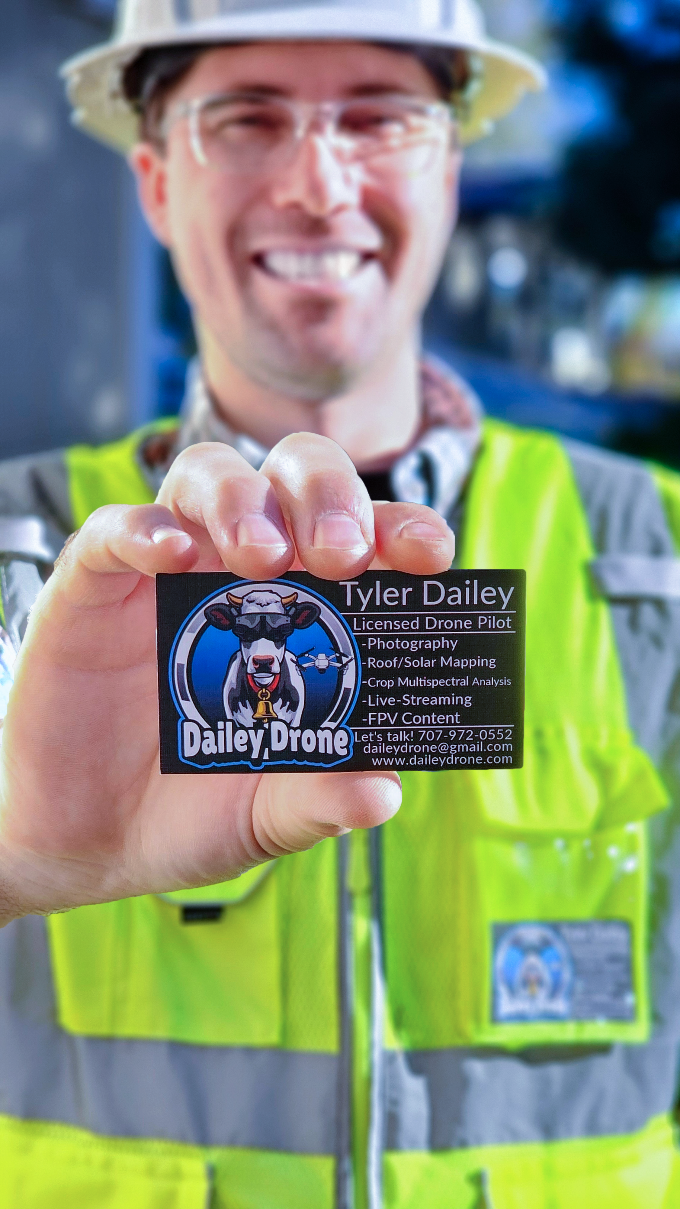A smiling man in a yellow safety vest and hard hat holding a business card for Dailey Drone, a licensed drone pilot company. The man is wearing glasses and standing outdoors.