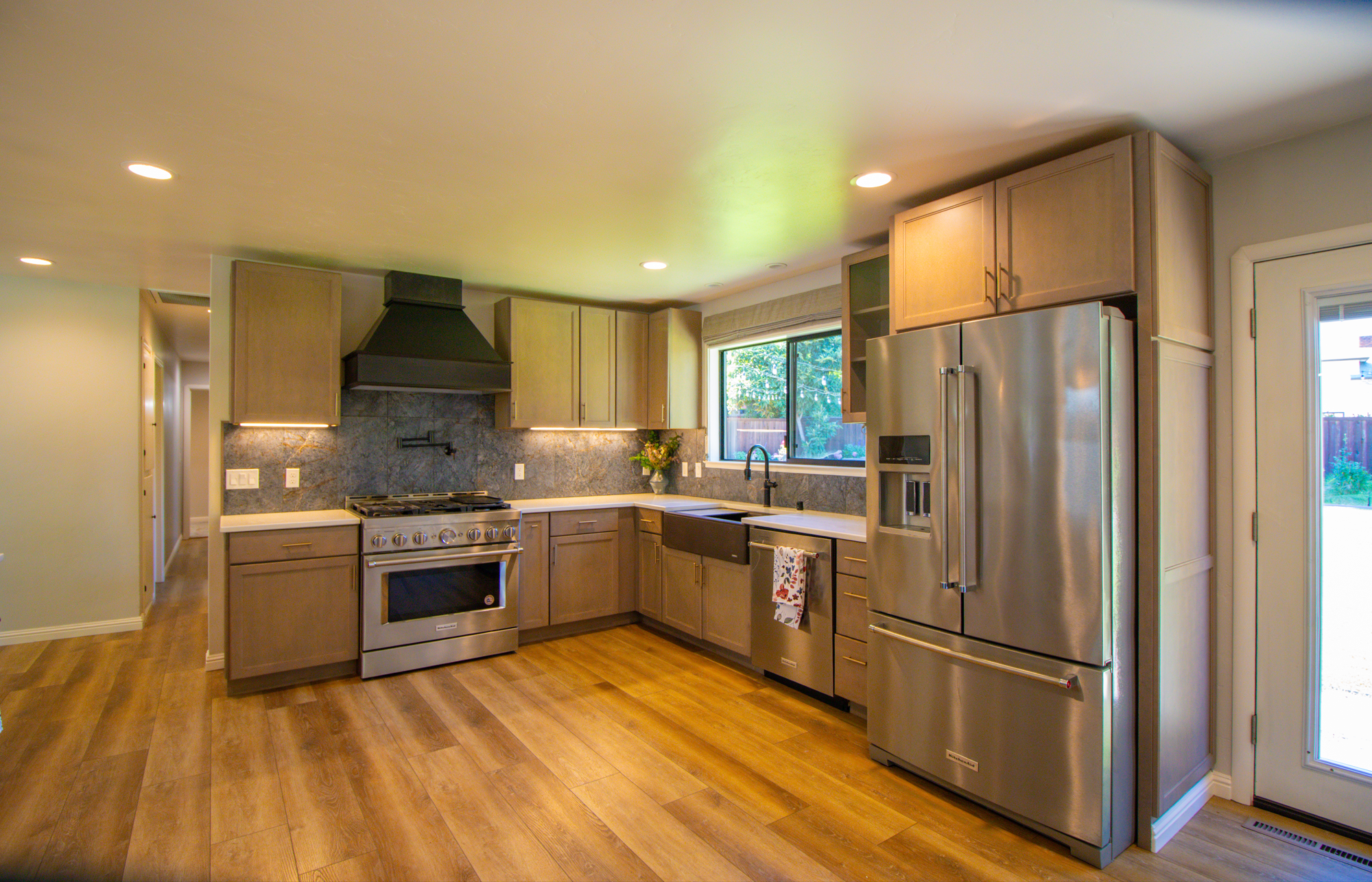 Kitchen with wooden cabinets, stainless steel refrigerator, oven, black range hood, and window overlooking a backyard with trees.