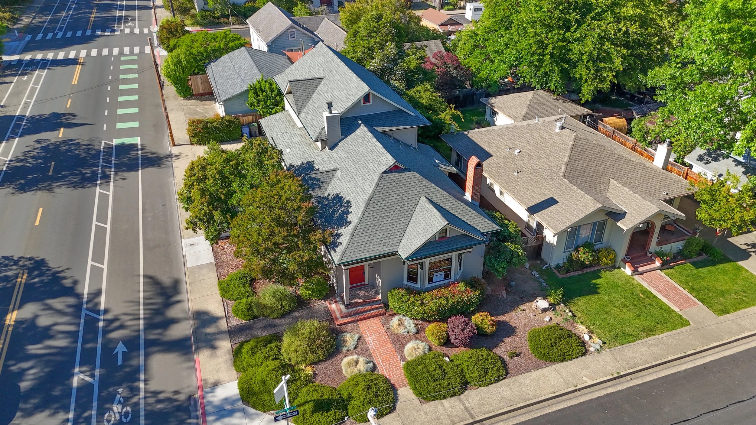 Aerial view of a residential neighborhood with several houses, trees, and a street with bike lanes and crosswalks.