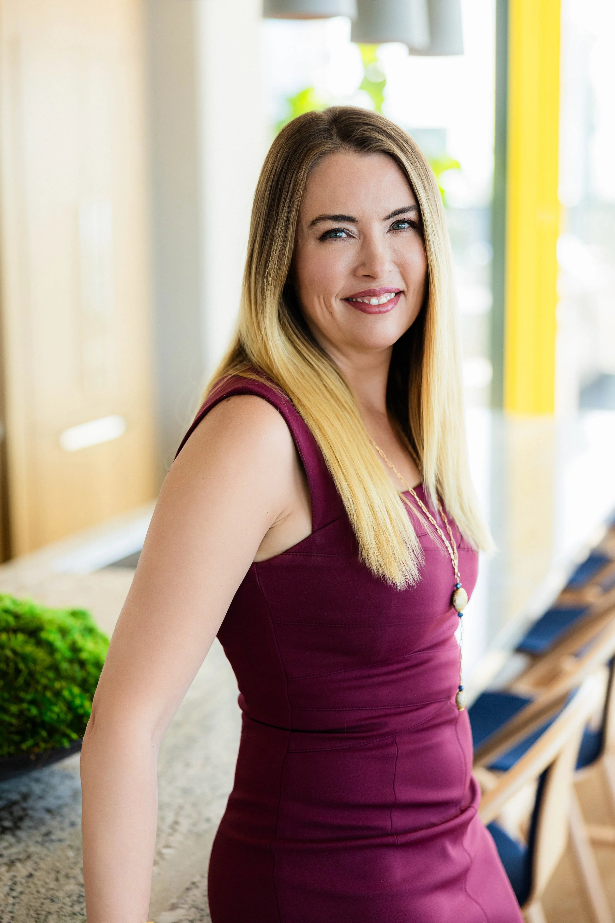A woman with long blonde hair, wearing a sleeveless burgundy dress and a necklace, smiling in a brightly lit indoor space.