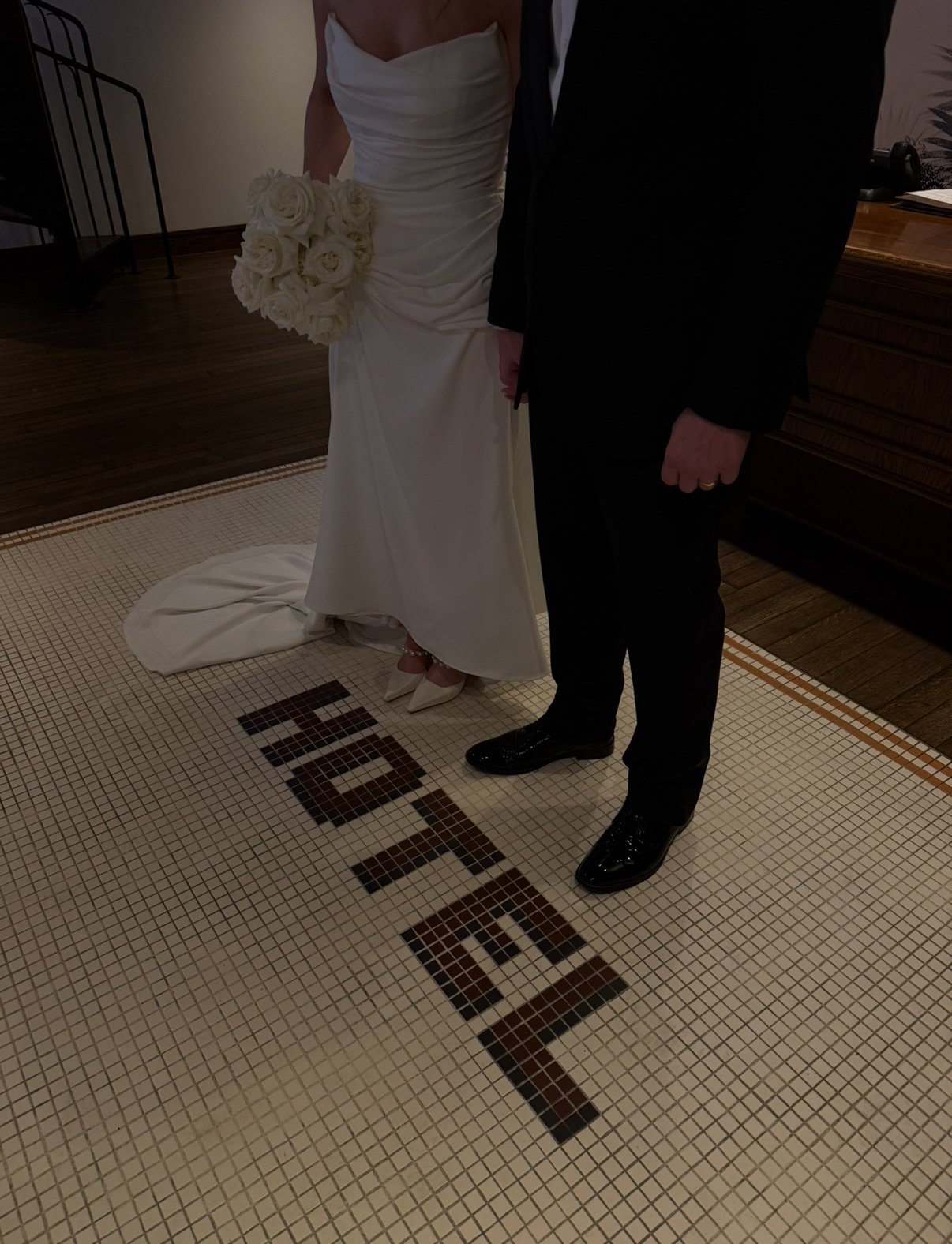 A bride in a white wedding gown holding a bouquet of white roses stands next to a groom in a black suit on mosaic tiles spelling "HOTEL."
