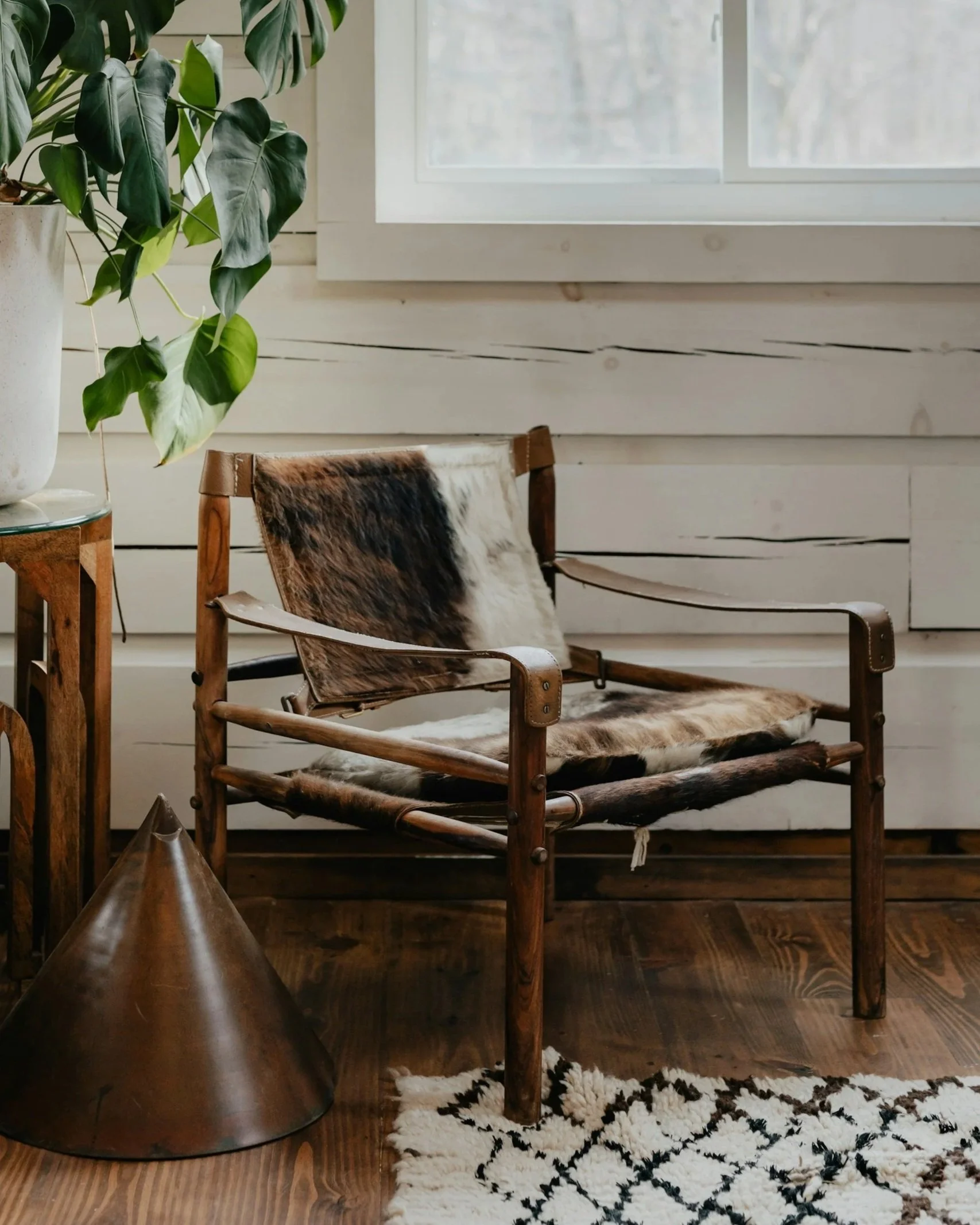 A cozy corner with a wooden chair draped with cowhide, a large potted plant with green leaves on a small table, a patterned rug on a wooden floor, and a window with white trim in the background.