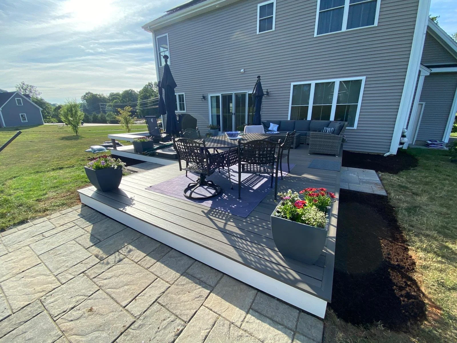 A backyard patio with outdoor furniture, including a dining table with chairs, a seating area with a sectional sofa, umbrellas, and potted flowers, attached to a two-story house with beige siding and large windows. The yard has a grassy area and a st