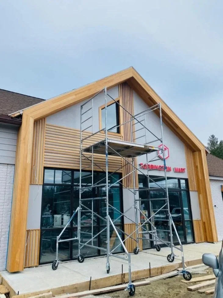 Construction scaffolding set up in front of a modern retail building with large glass windows and wood paneling under a cloudy sky.