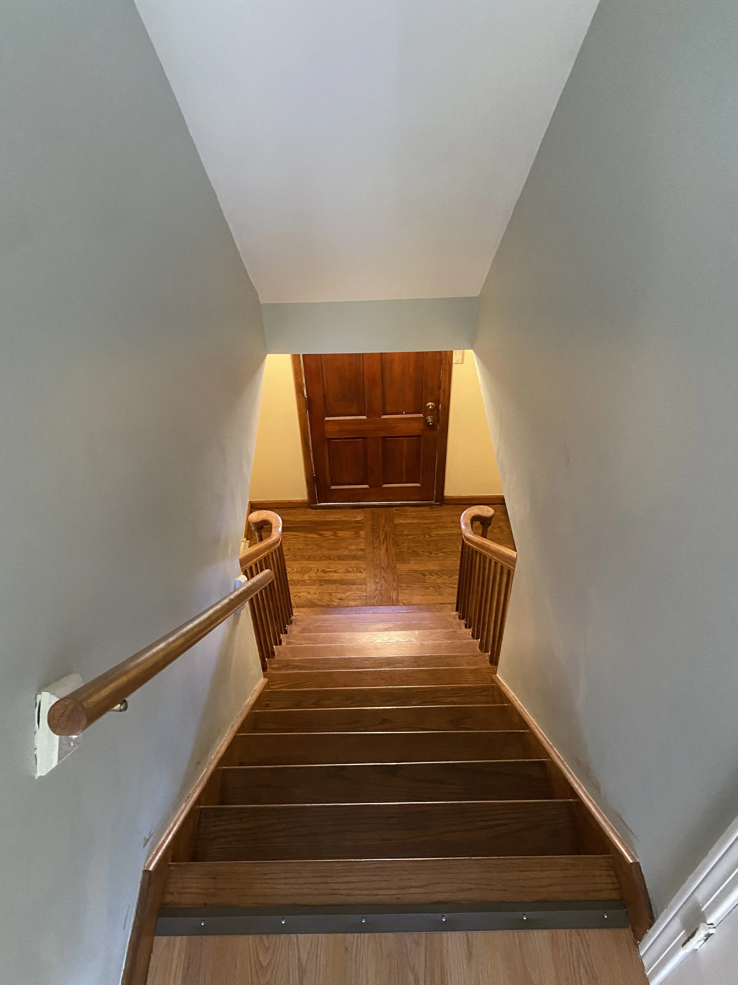 Wooden staircase leading down to a closed wooden door at the bottom with a brass doorknob. Light green walls and a wooden handrail on the left side.