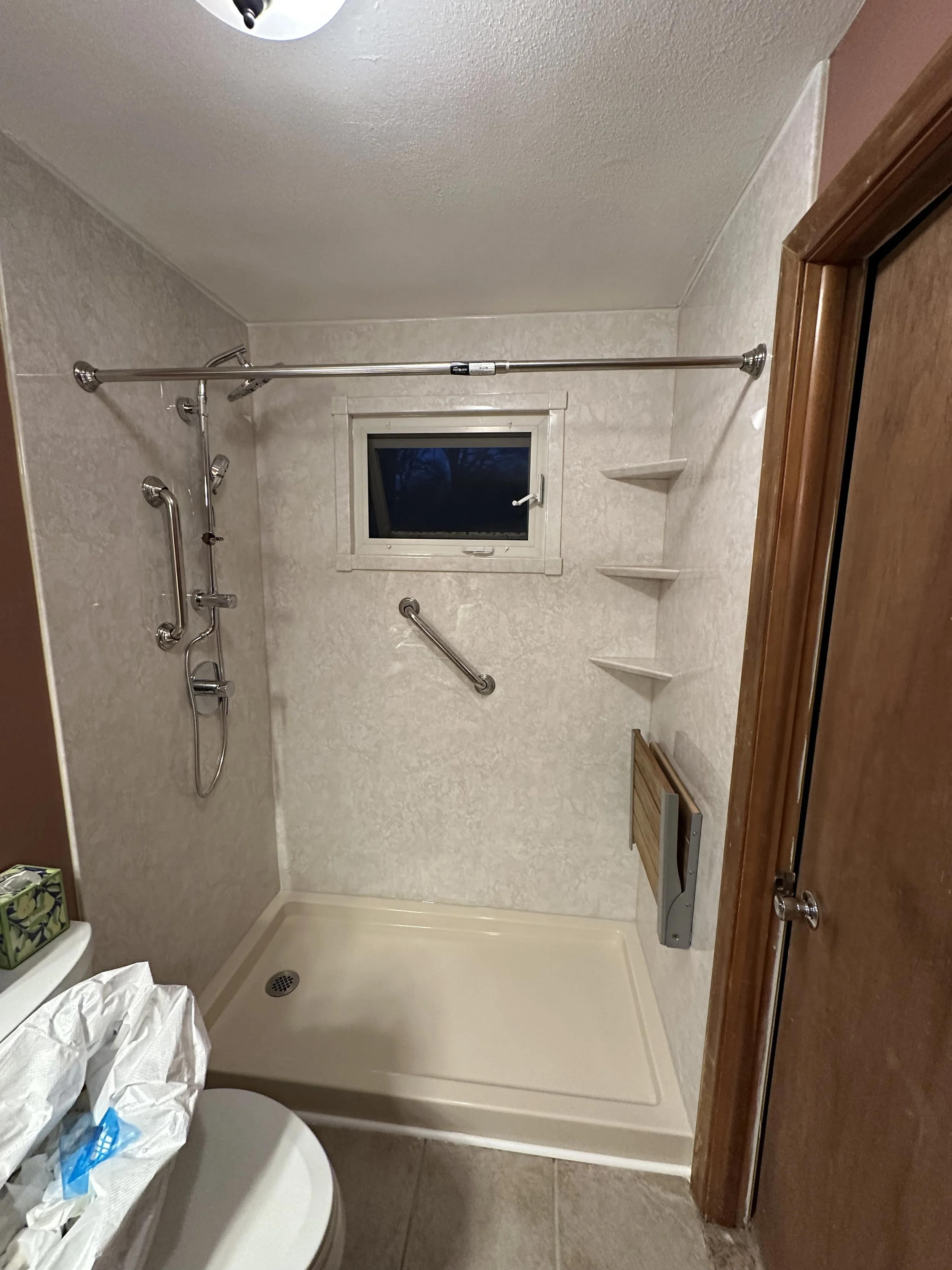 Empty walk-in shower with beige walls, a small window, built-in corner shelves, a grab bar, and a handheld showerhead with multiple connectors. A toilet with a tissue box is partially visible in the foreground, and a wooden door is on the right side.