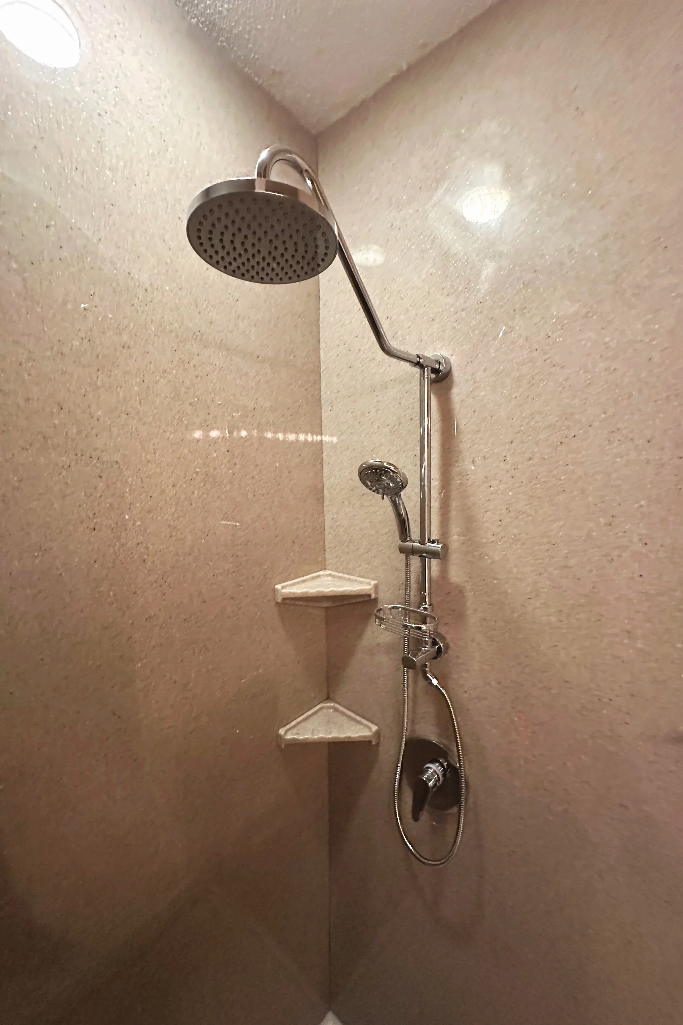 Shower with beige walls, a large overhead showerhead, a smaller handheld showerhead, a soap dish, and a water control handle.