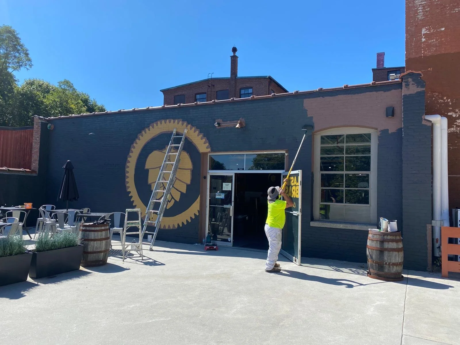 A man in a neon yellow shirt and white pants is painting the exterior wall of a building. He is holding a long paint roller and standing in front of the open door. The building has a large circular logo painted on the wall that looks like a gear with