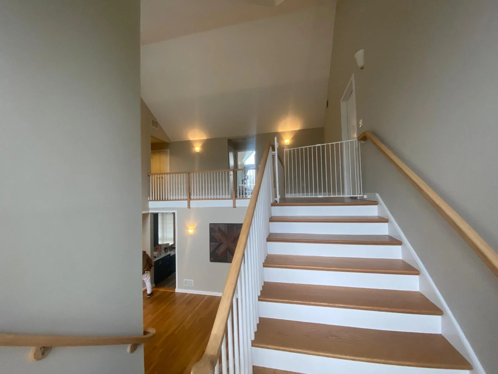 Interior of a modern home with a staircase leading to an upper floor, featuring wooden handrails and white risers, beige walls, and a lofted area with a railing. Warm lighting fixtures illuminate the space.