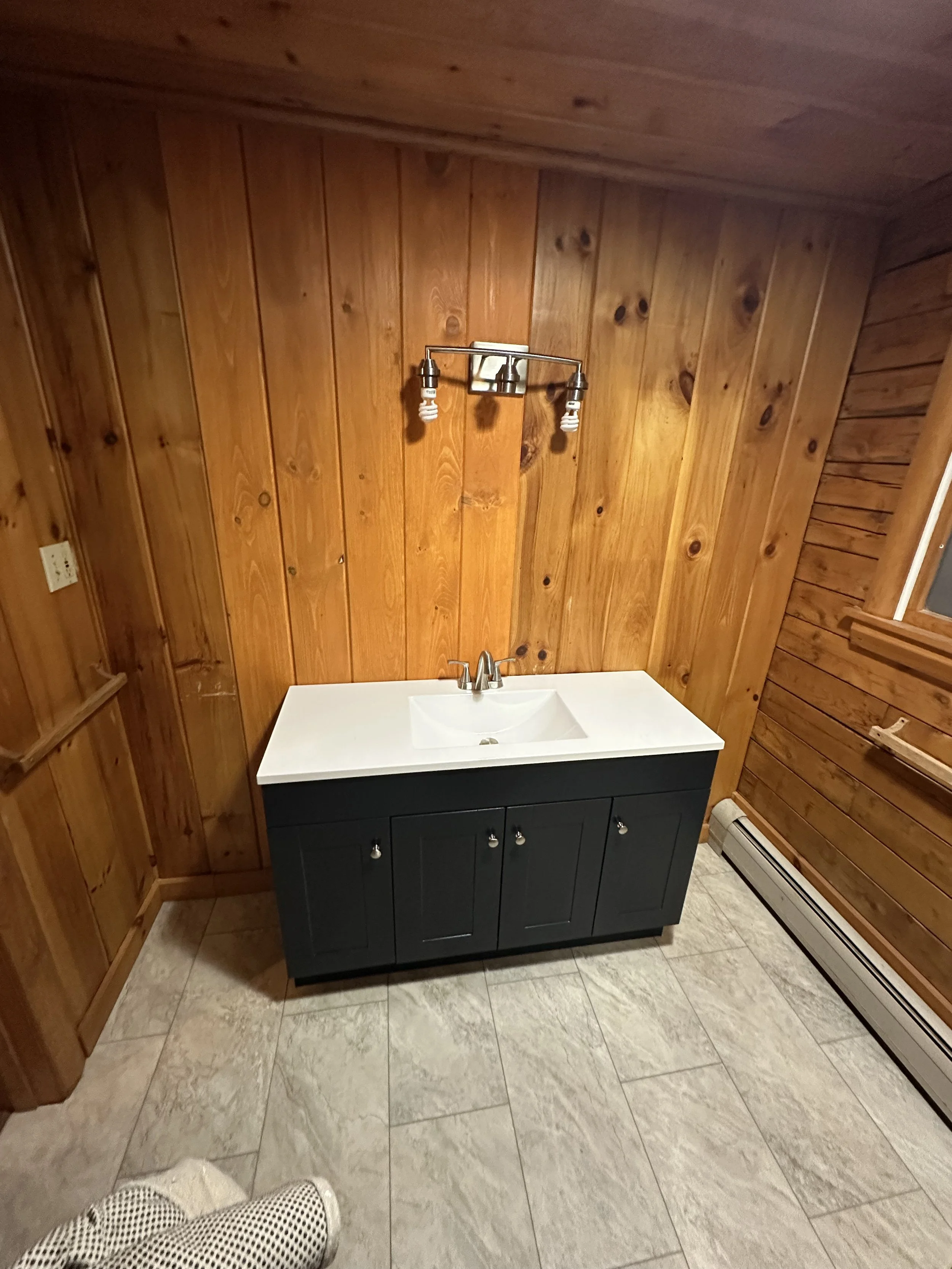 Bathroom with wooden-paneled walls, a dark blue vanity with white sink, a faucet, and two light fixtures above the sink, with a baseboard heater below the window on the right side.