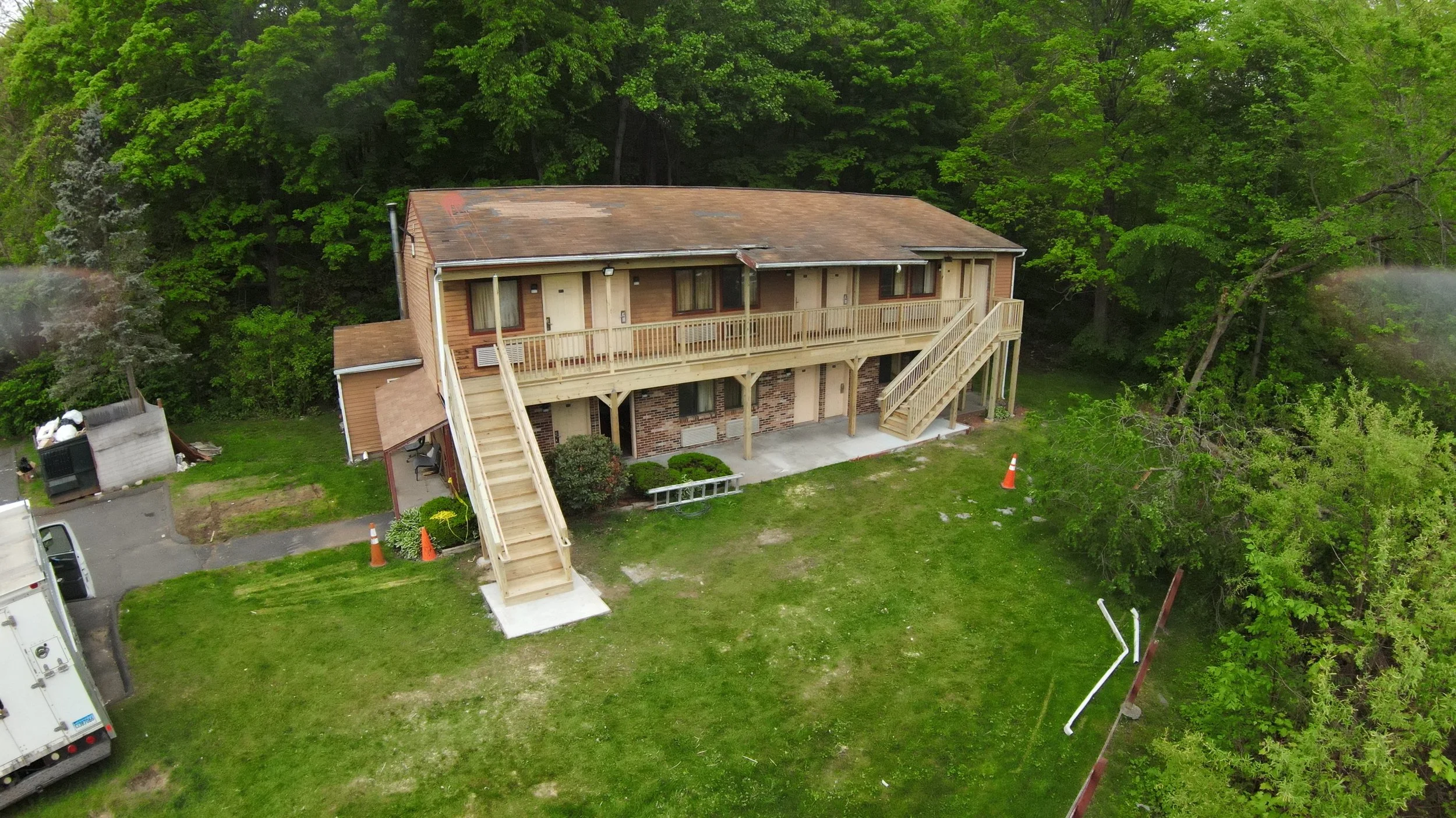 An aerial view of a two-story residential building with a wooden exterior, deck, and stairs, surrounded by green trees and grass.