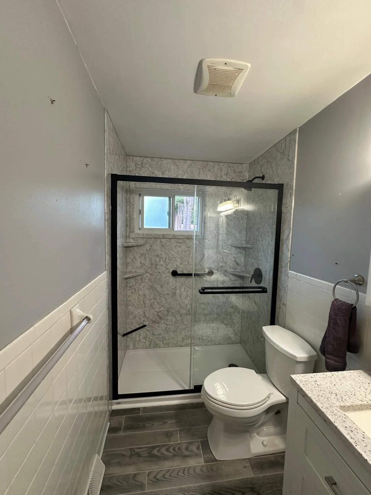 Bathroom with a glass enclosed shower, white toilet, granite countertop, gray wood-look tile flooring, gray painted walls, a small window, and a towel hanging on the wall.