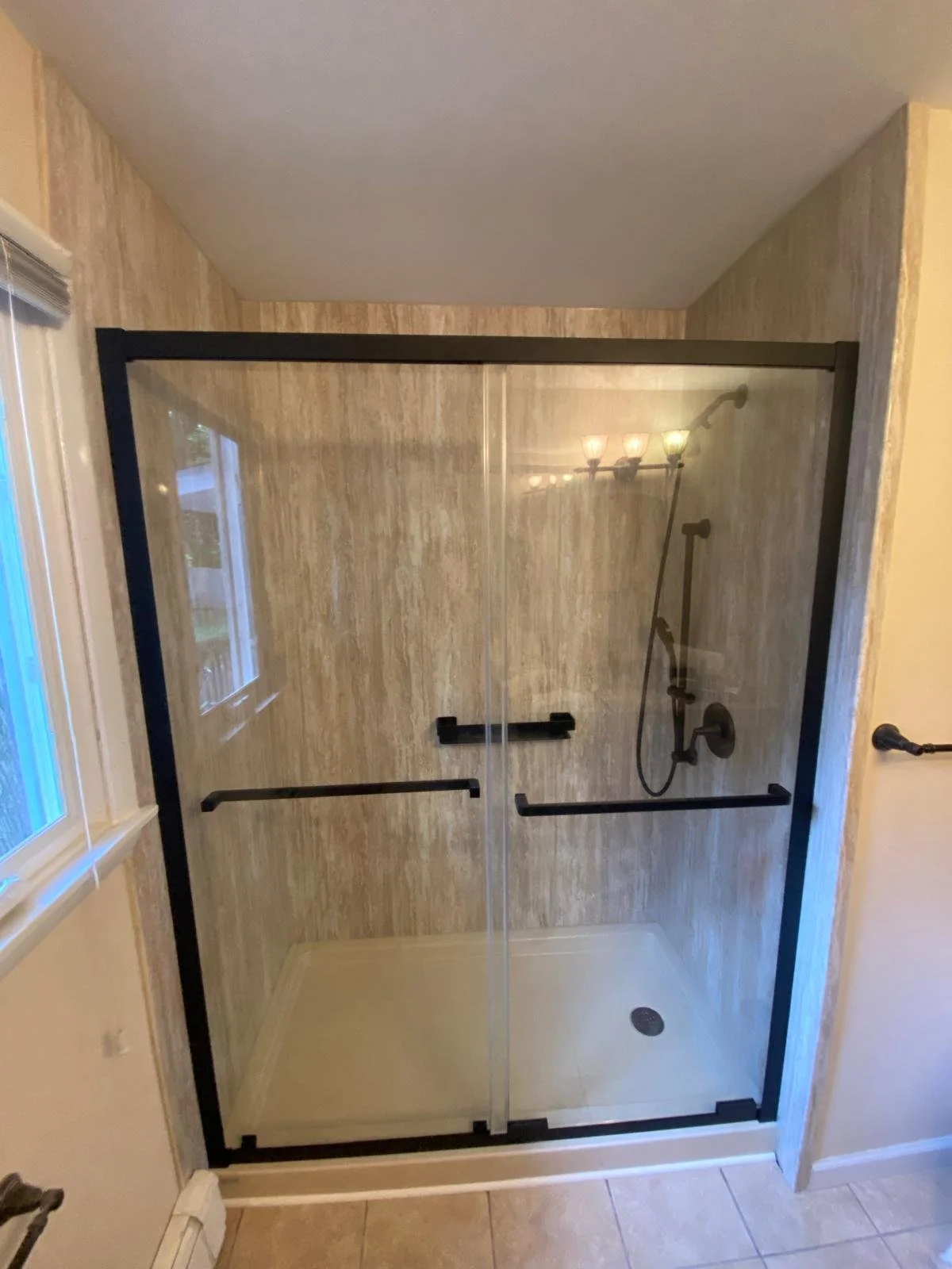 A walk-in shower with glass doors, beige marble walls, black grab bars, a black showerhead, and a handheld shower wand, with tiled flooring.