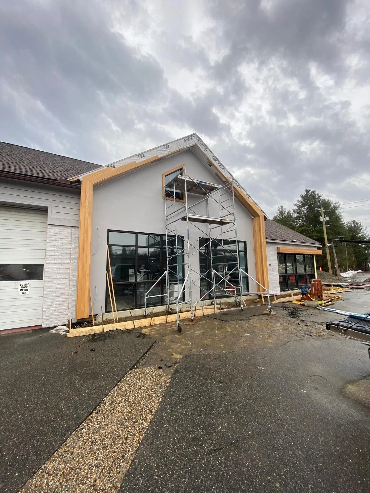 Building under construction with scaffolding, gray exterior walls, large glass windows, and a partially completed roof, clouds in the sky.