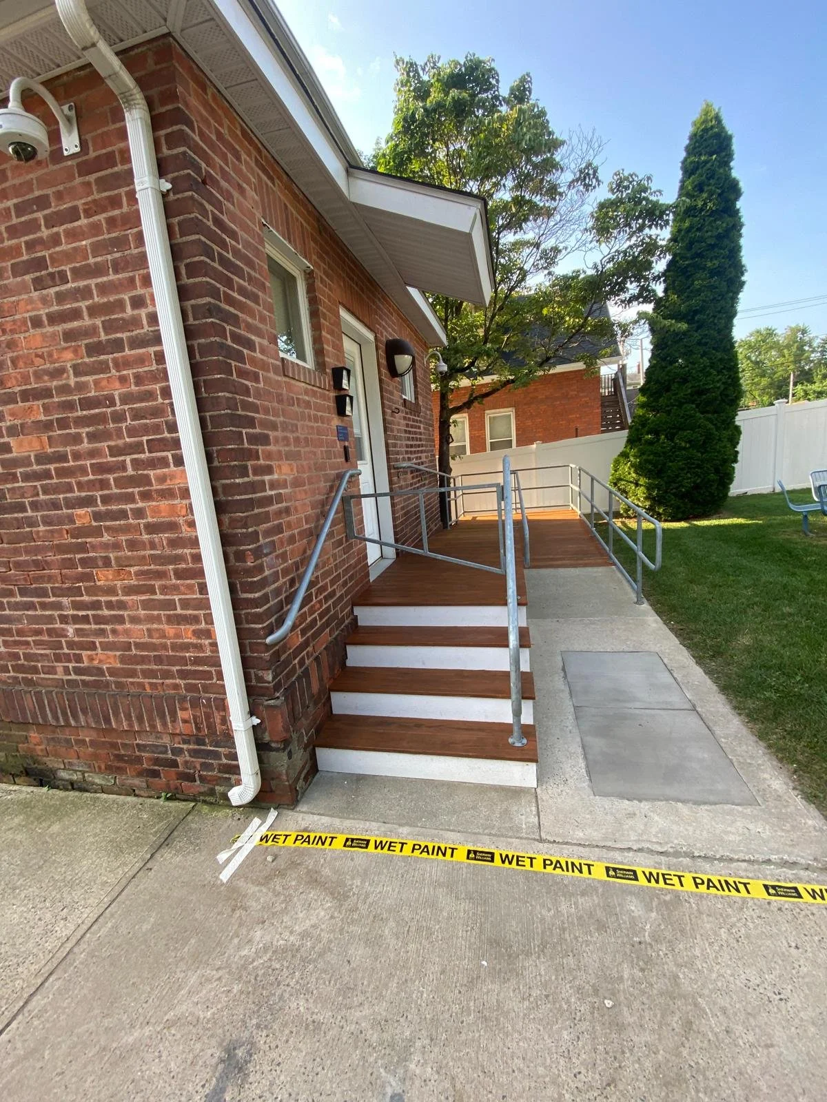 Exterior view of a brick building with a ramp and staircase for accessibility. Wet paint caution tape is on the sidewalk, trees and a white fence are in the background.