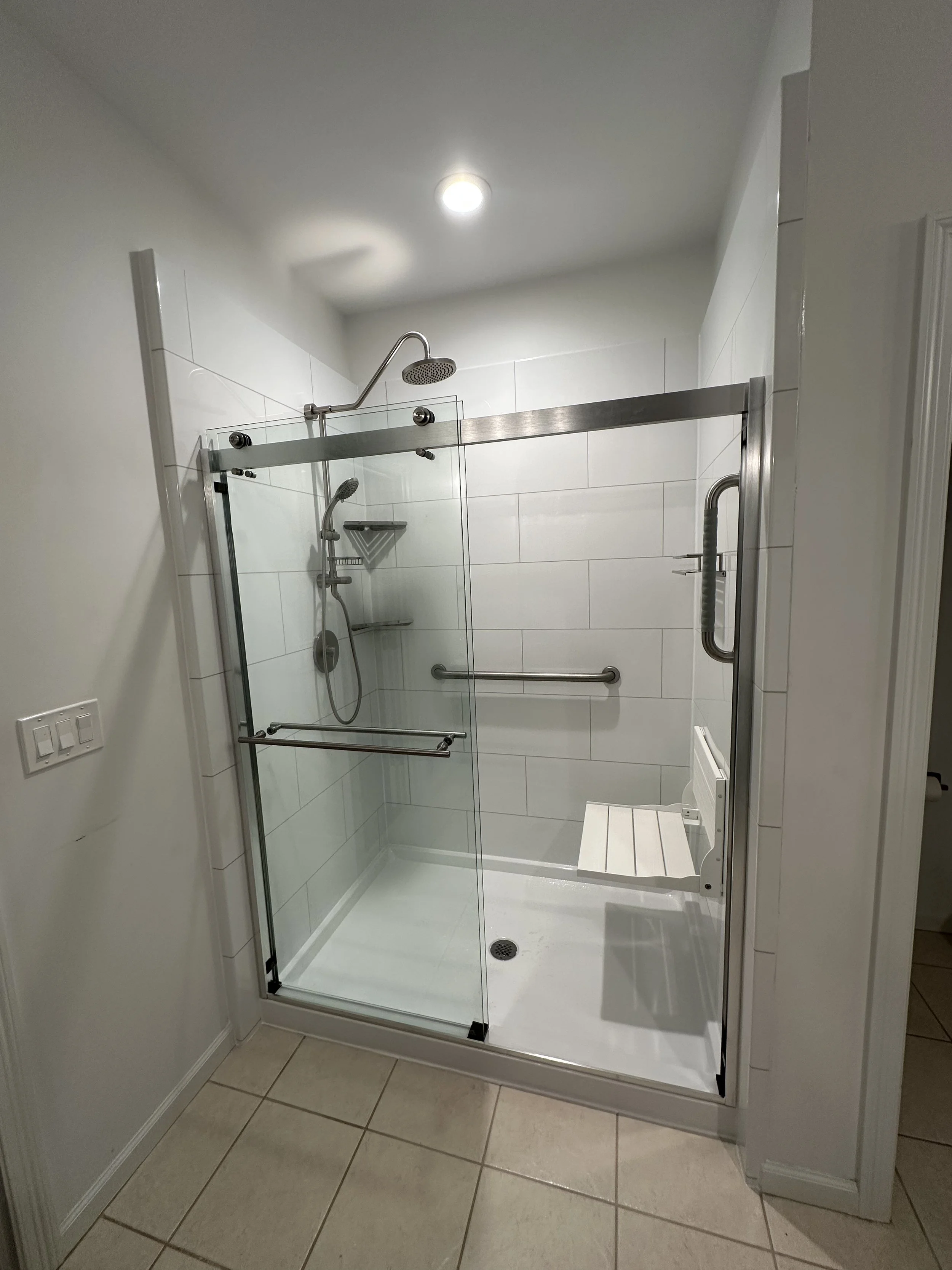 Clean walk-in shower with glass door, white tiled walls, overhead showerhead, handheld shower, a corner shelf, and a built-in bench inside a bathroom.