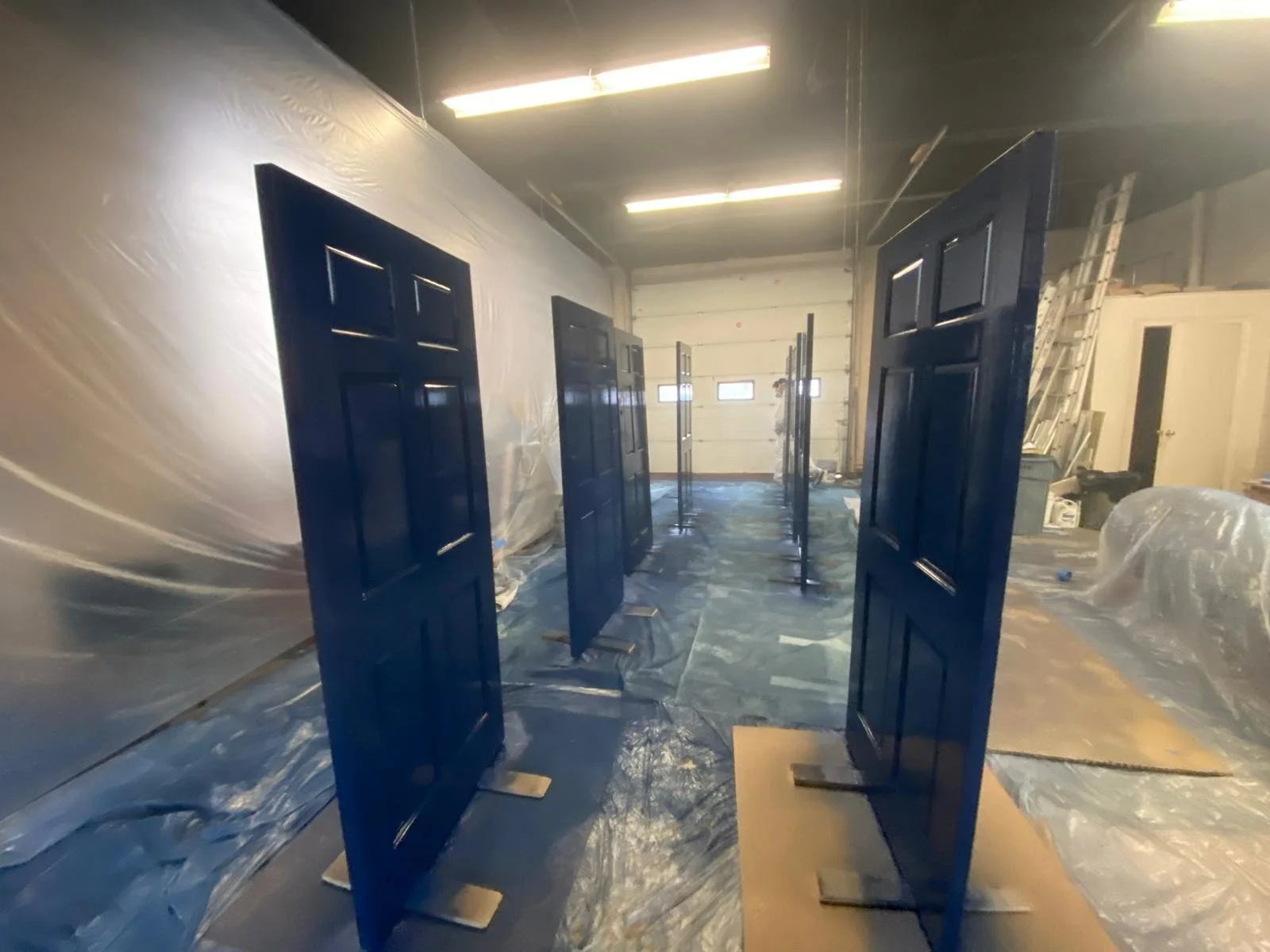Several blue doors standing upright in a warehouse or workshop with plastic sheeting on the floor and walls, and fluorescent lights overhead.
