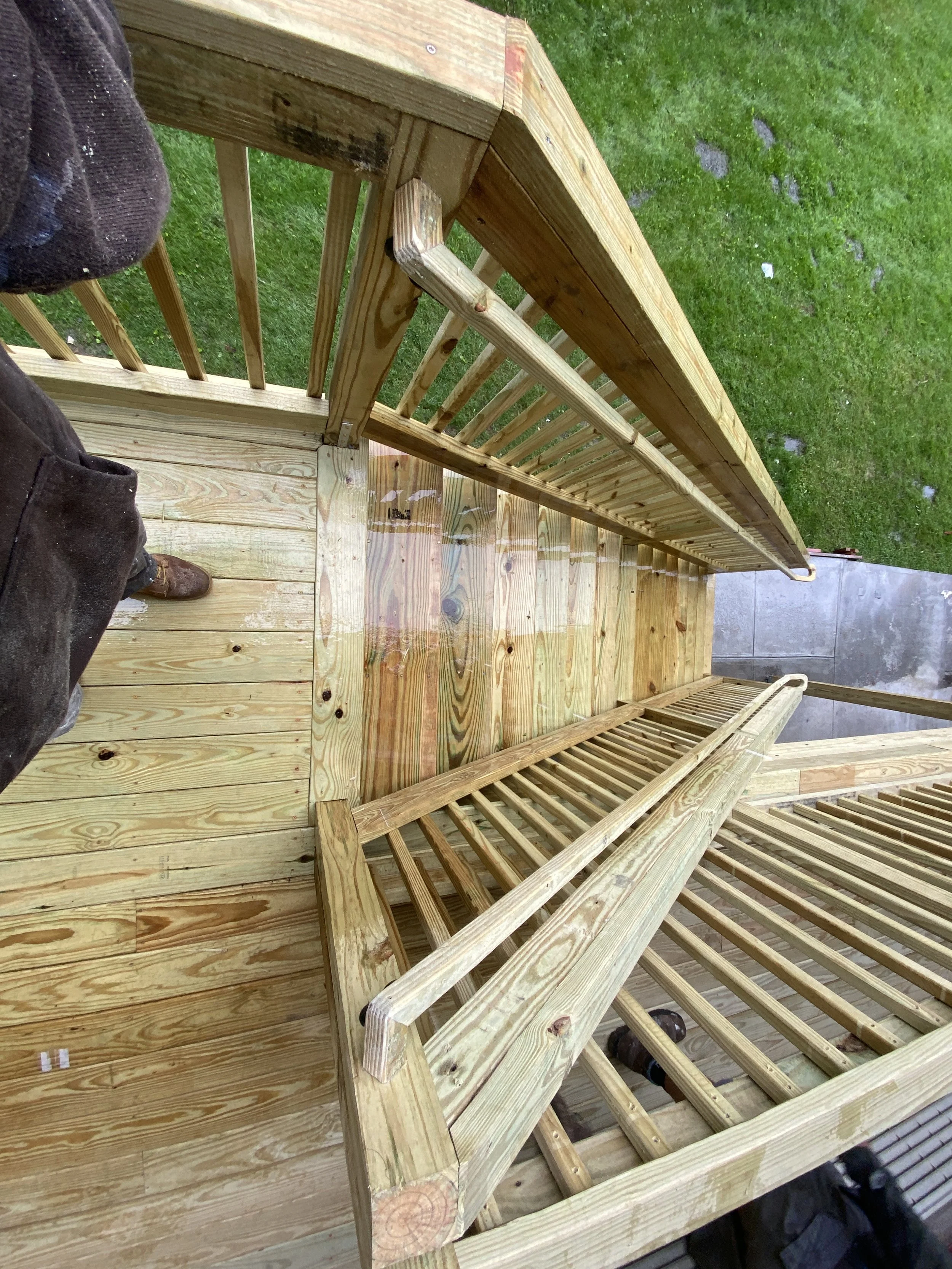 A wooden deck with a railing, taken from above, showing wet wood and a grassy yard in the background.