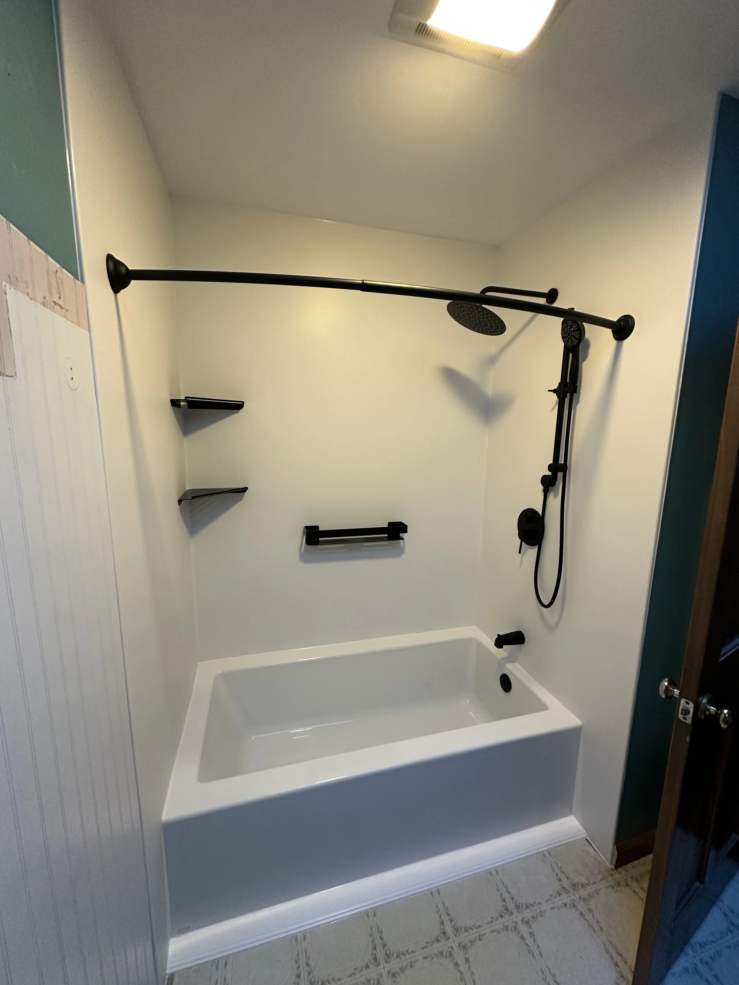 Small bathroom with a white bathtub, black shower fixtures, and a wall-mounted showerhead with a black pipe. Two black shelves and a towel bar are on the wall, and the floor has patterned tiles.