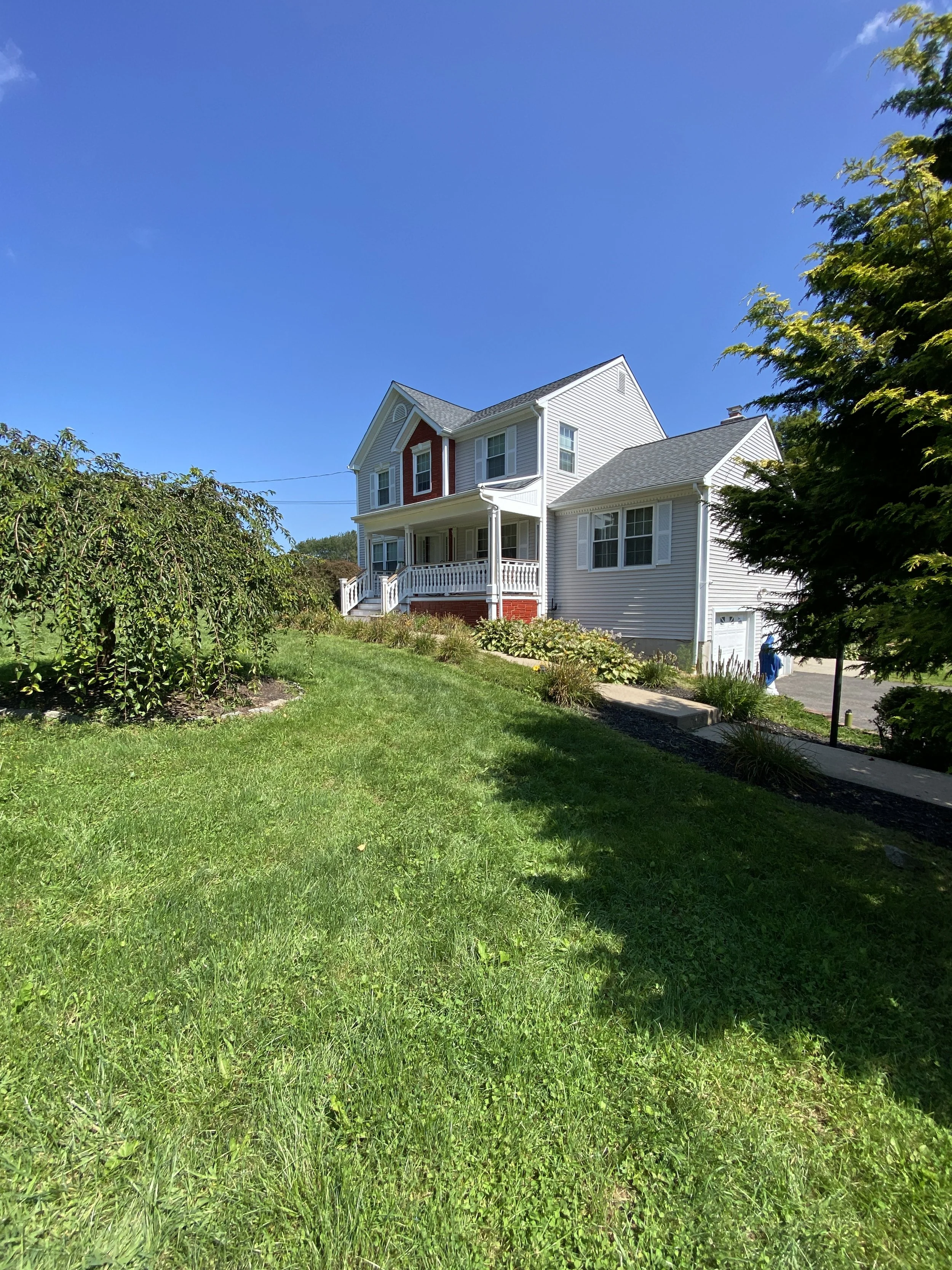 A two-story white house with a front porch, surrounded by green grass, trees, and shrubs, under a clear blue sky.
