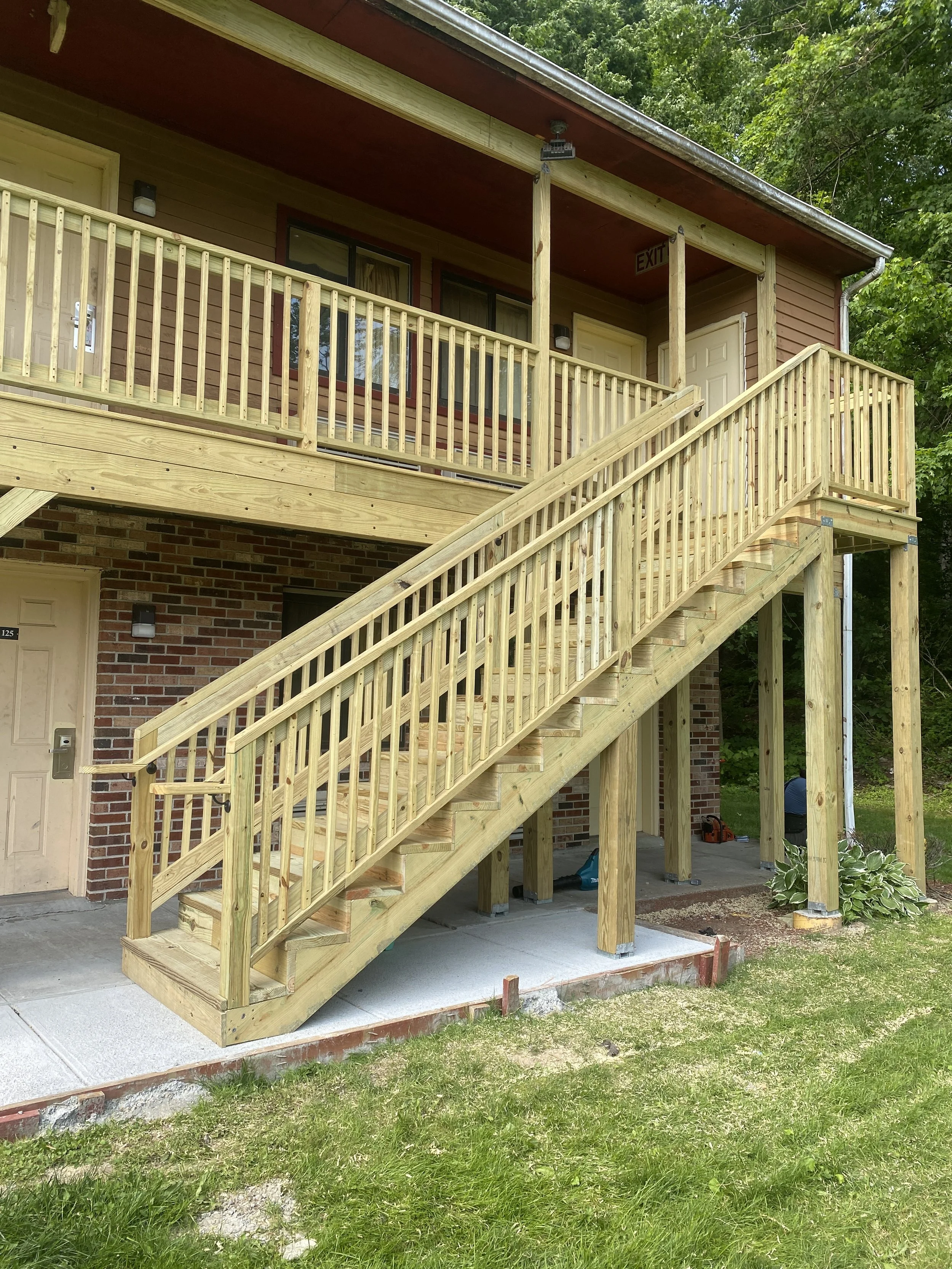 Newly constructed outdoor wooden staircase with handrails leading from ground level to an upper balcony of a brick and siding residential building, surrounded by green trees.