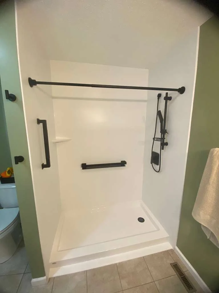 Empty walk-in shower with white walls, black grab bars, a showerhead with a hose, and a corner shelf, next to a green bathroom wall and tile floor.