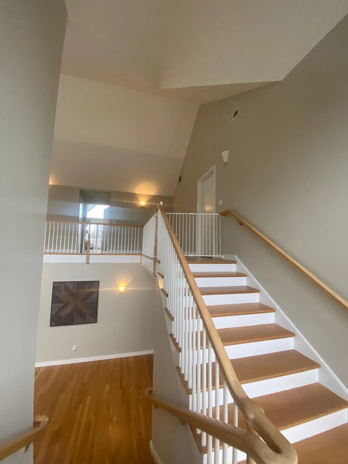 Interior view of a staircase in a modern home with wooden handrails, white balusters, beige walls, hardwood flooring, and wall-mounted light fixtures.