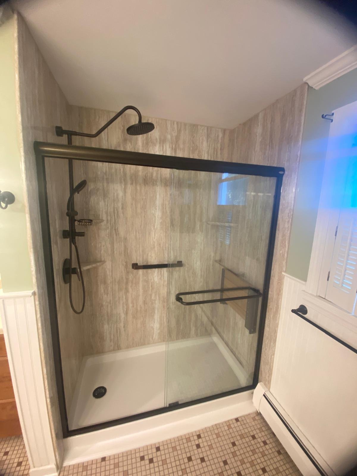 A bathroom with a walk-in shower enclosed by a glass door, featuring black fixtures including a showerhead and a handheld shower. The shower walls are tiled with beige, textured tiles and there is a small built-in shelf for toiletries. The bathroom f