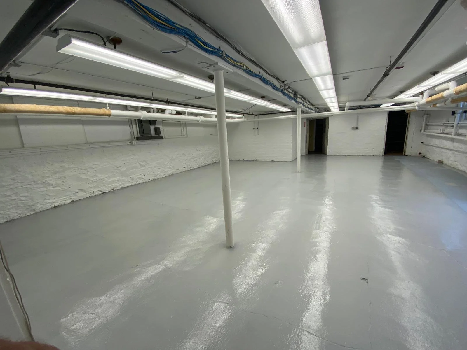 Empty basement with white painted brick walls, exposed ceiling with pipes and wiring, and a shiny concrete floor.