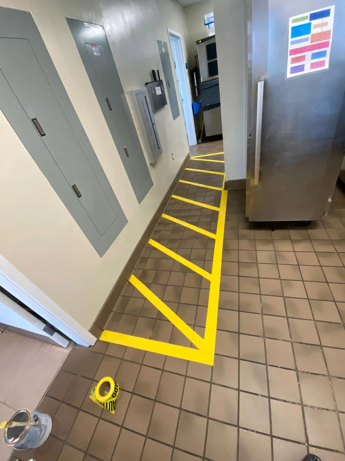 A tiled floor with bright yellow diagonal and striped caution tape marking a small area near a wall with electrical panels and a doorway, in a commercial kitchen or utility room.