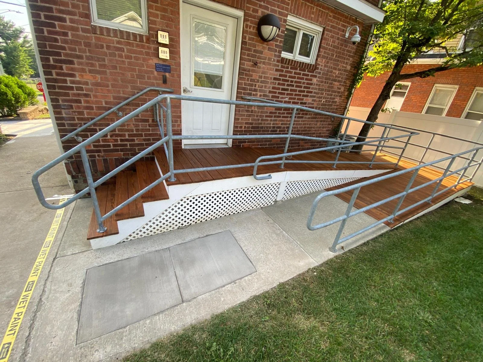 A brick building entrance with a white door and two small windows, a black outdoor light, and a clean, newly built wooden wheelchair ramp with metal railings.