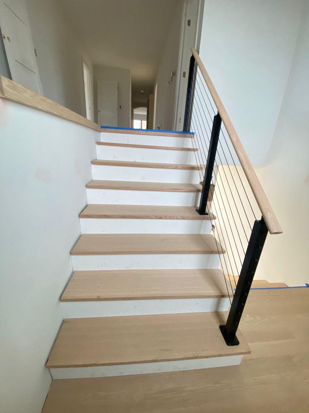 Interior view of a staircase under construction with light wooden steps, black metal railing with horizontal wires, and white walls.