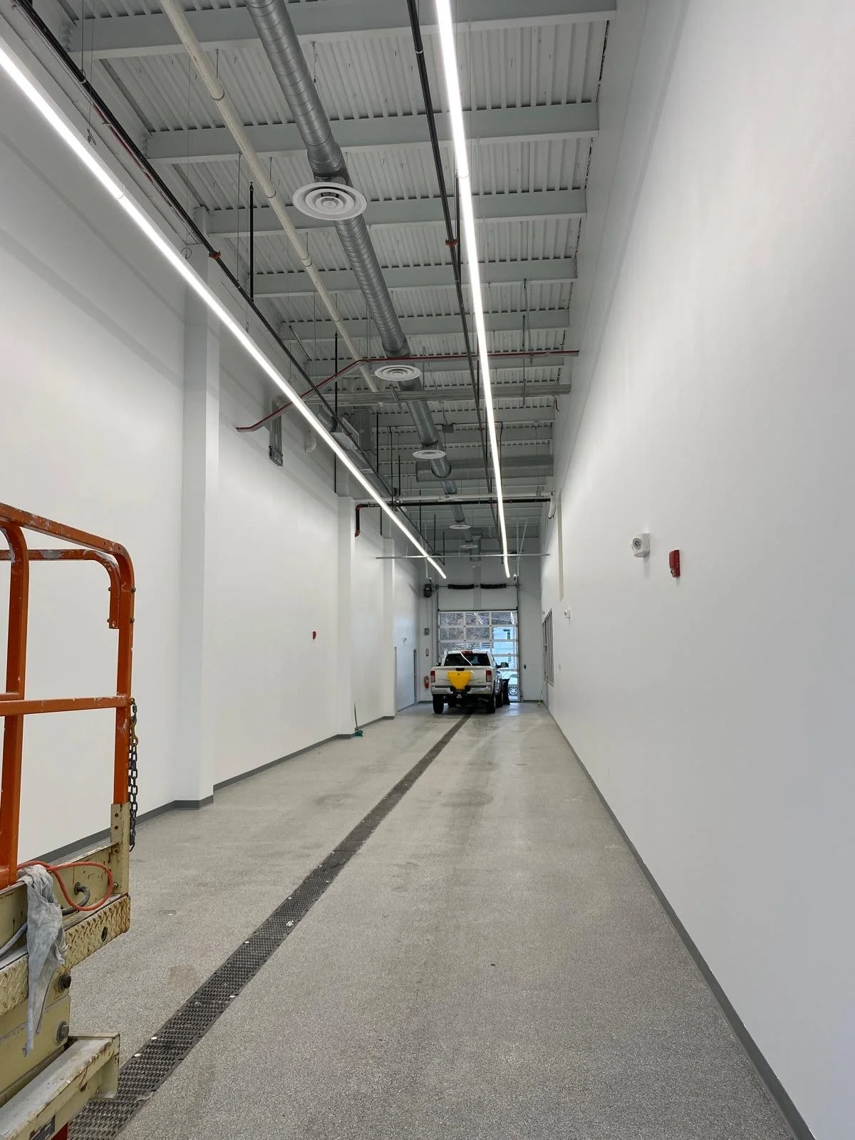 Empty garage with white walls, gray flooring, and a white pickup truck parked near the back, with construction equipment on the left side.