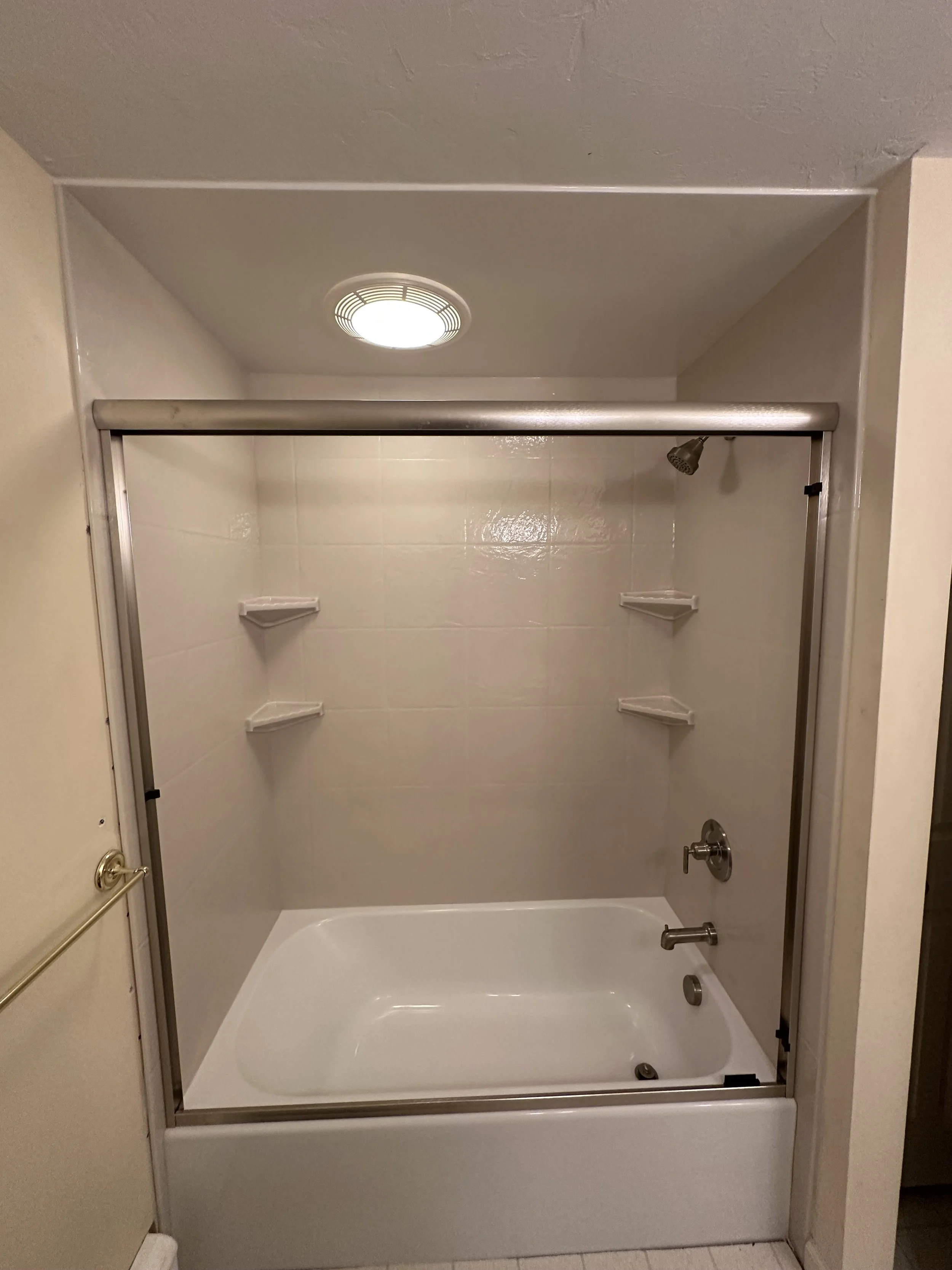 A white bathtub with a sliding glass door, mounted showerhead, and built-in corner shelves against a tiled wall in a bathroom.