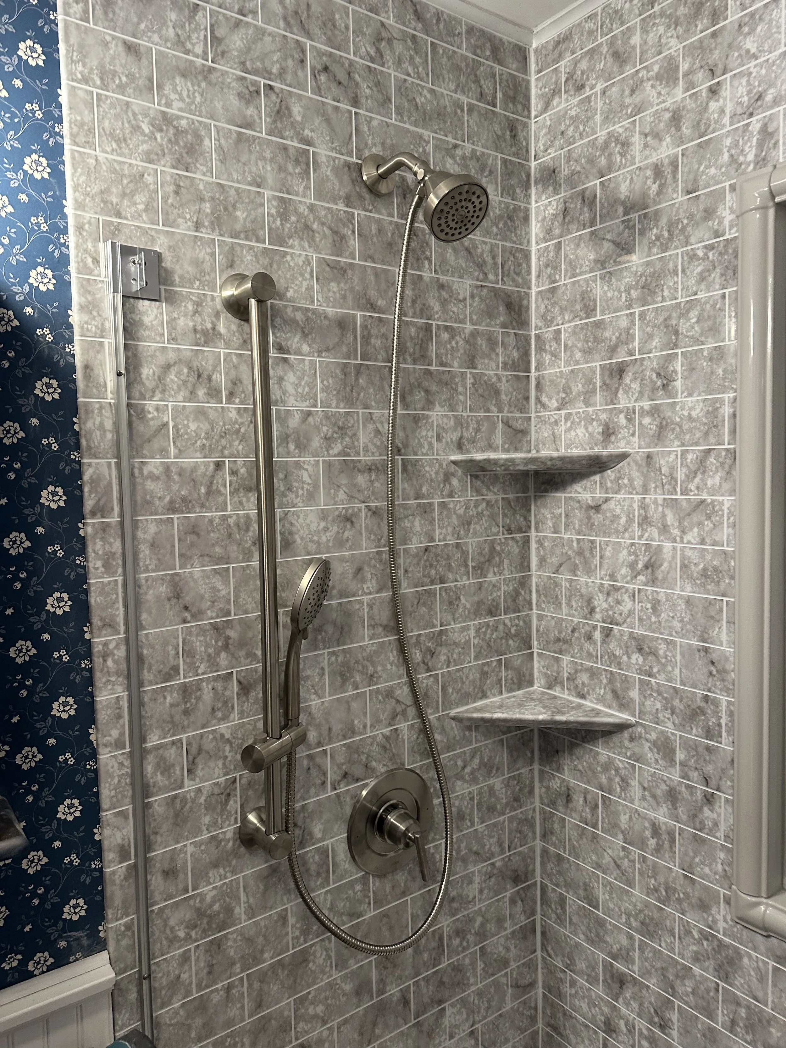 Gray tiled shower with a metal handheld showerhead, a curved metal pipe showerhead, and built-in corner shelves.