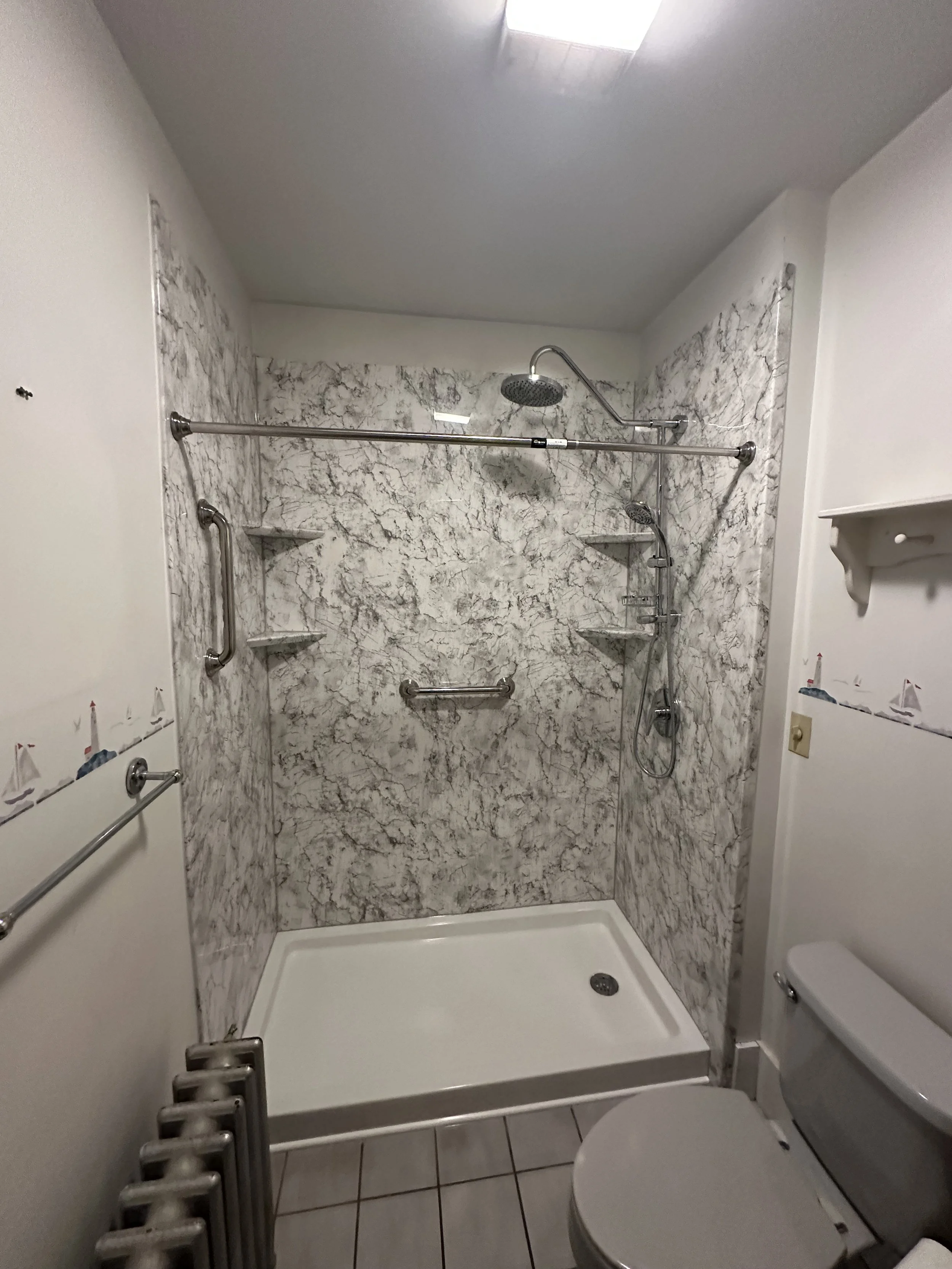 A modern bathroom with a marble shower area, two small wall shelves, grab bars, and a handheld showerhead. The bathroom has tiled flooring and a toilet next to the shower. There is a ceiling light above.
